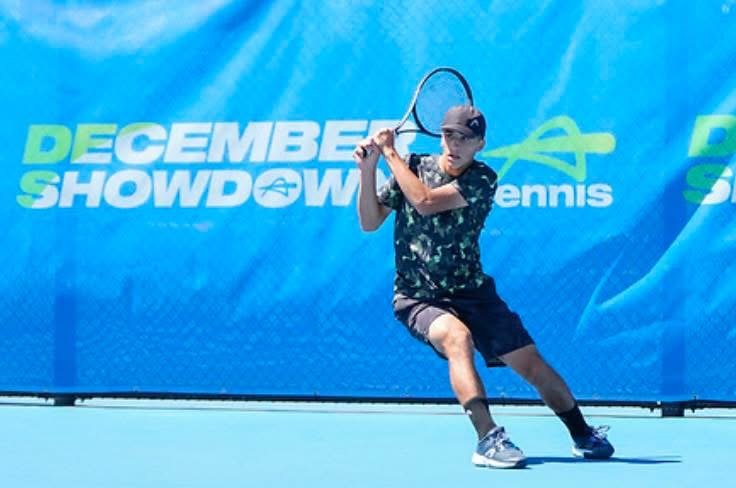 Young boy playing tennis on an outdoor court with a blue backdrop that says 'December Showdown Tennis'.