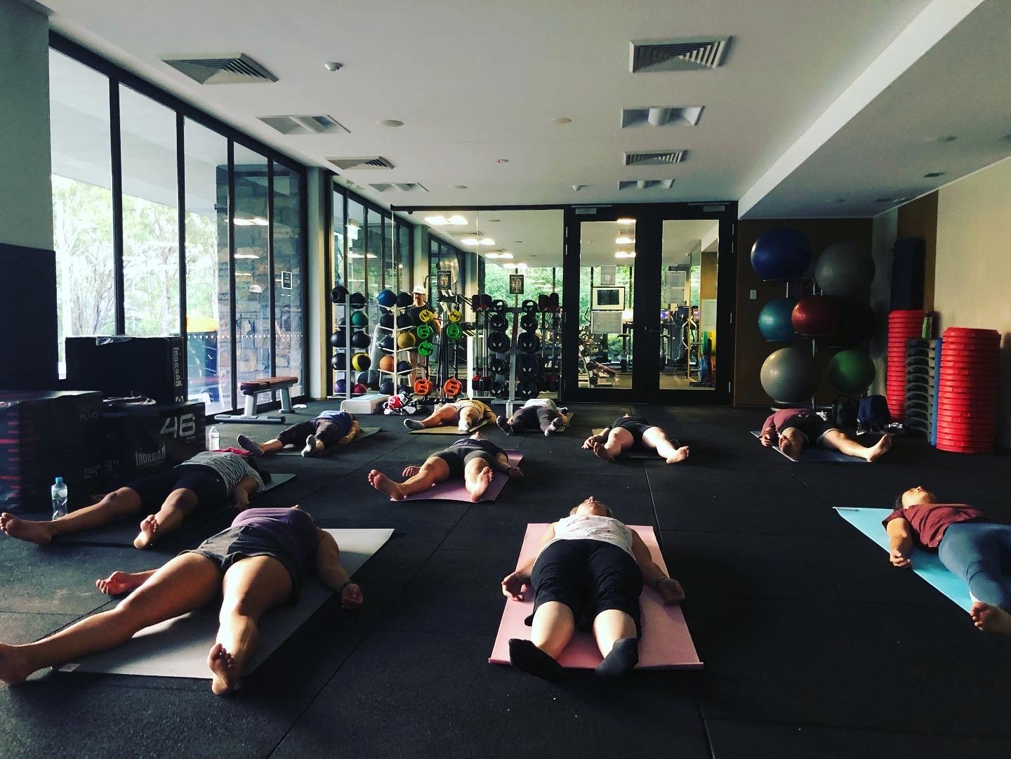 Group of people practicing yoga on mats in a gym with large windows and exercise equipment in the background.