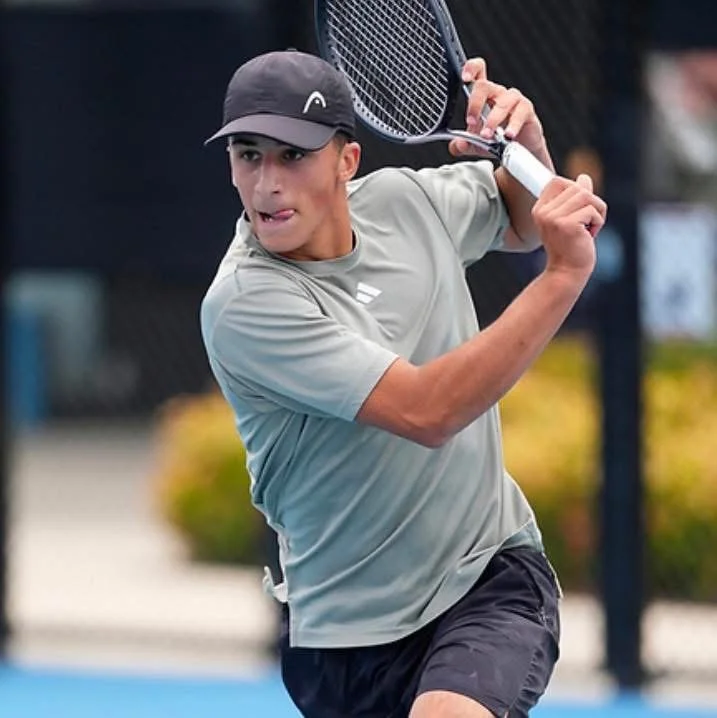A young male tennis player wearing a black cap and grey t-shirt prepares to hit a tennis ball with a backhand stroke on an outdoor court.