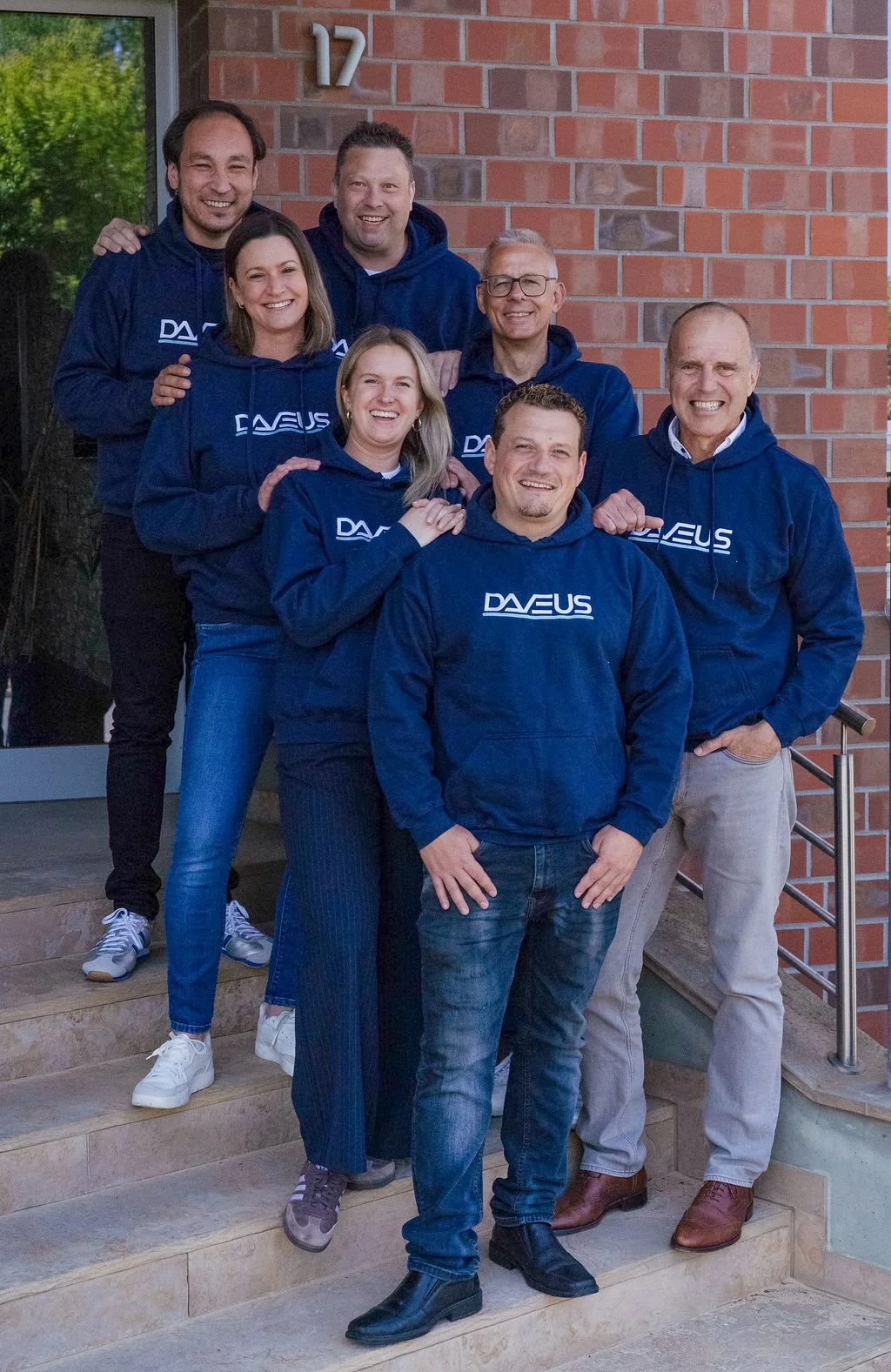 Das Bild zeigt die Mitarbeiterinnen und Mitarbeiter der Firma Daveus. Sie tragen blaue Hoodies mit dem Firmenlogo und lächeln in die Kamera.
