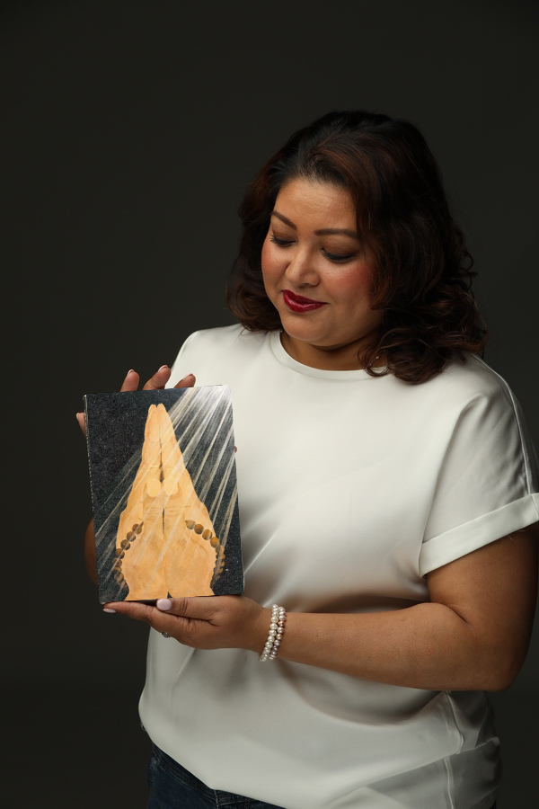 Vrouw met donkerbruin haar die een foto vasthoudt van een plaquette met beelden van een asymmetrische kunstwerk, tegen een donkere achtergrond.