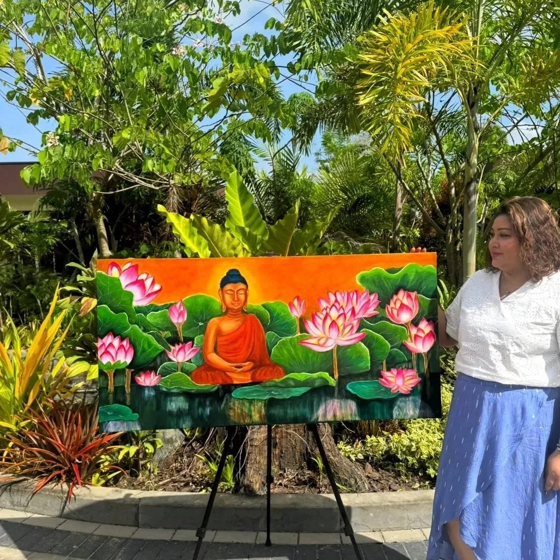 Een vrouw staat naast een kleurrijk schilderij van een boeddha die in een lotus-veld zit, omringd door grote roze lotusbladeren en waterplanten, buiten in een tuin met groene bomen en een blauwe lucht.