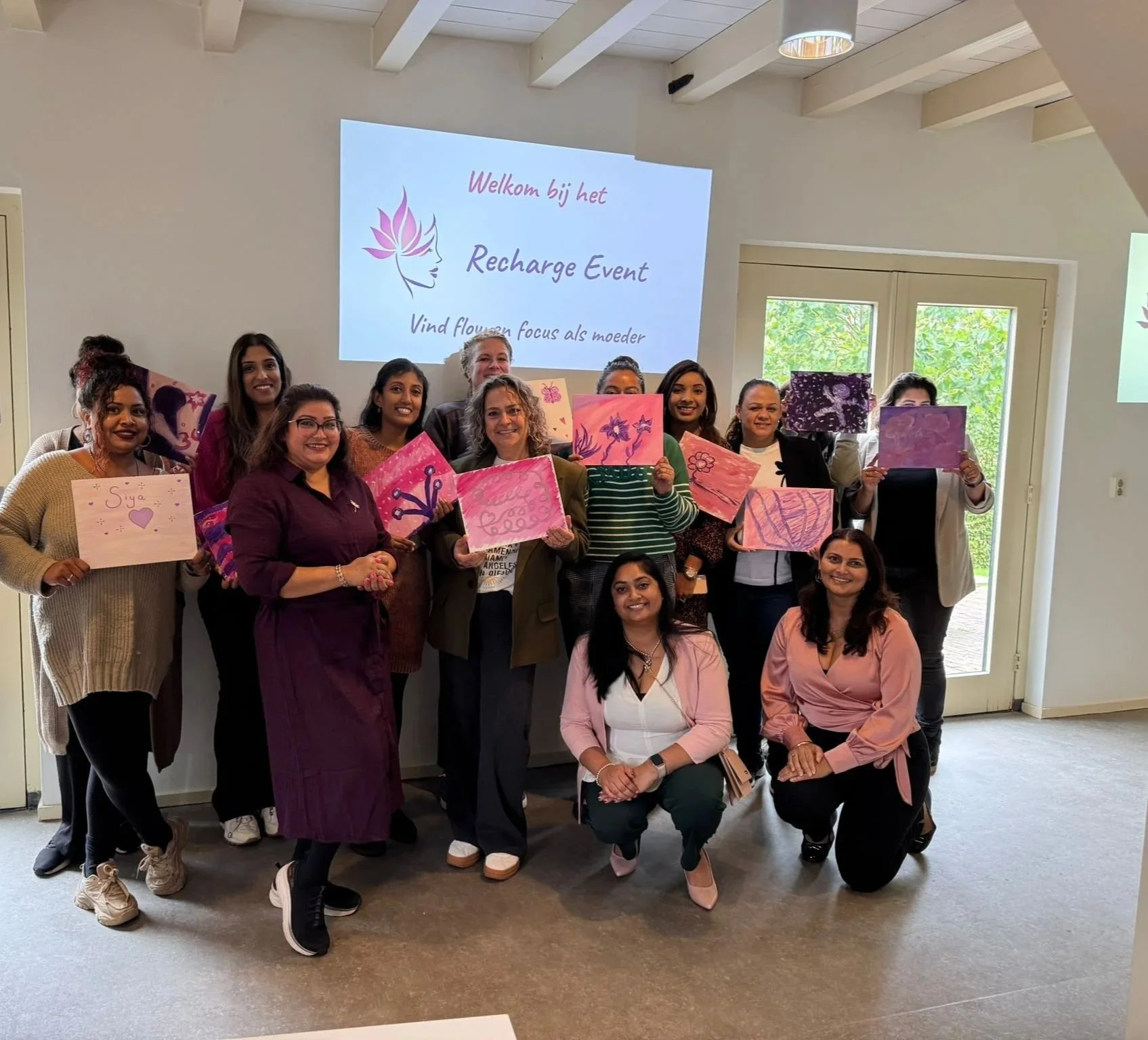 Groep vrouwen die tijdens een workshop of evenement een blad met kleurrijke schilderijen vasthouden, voor een whiteboard met een welkomstboodschap.