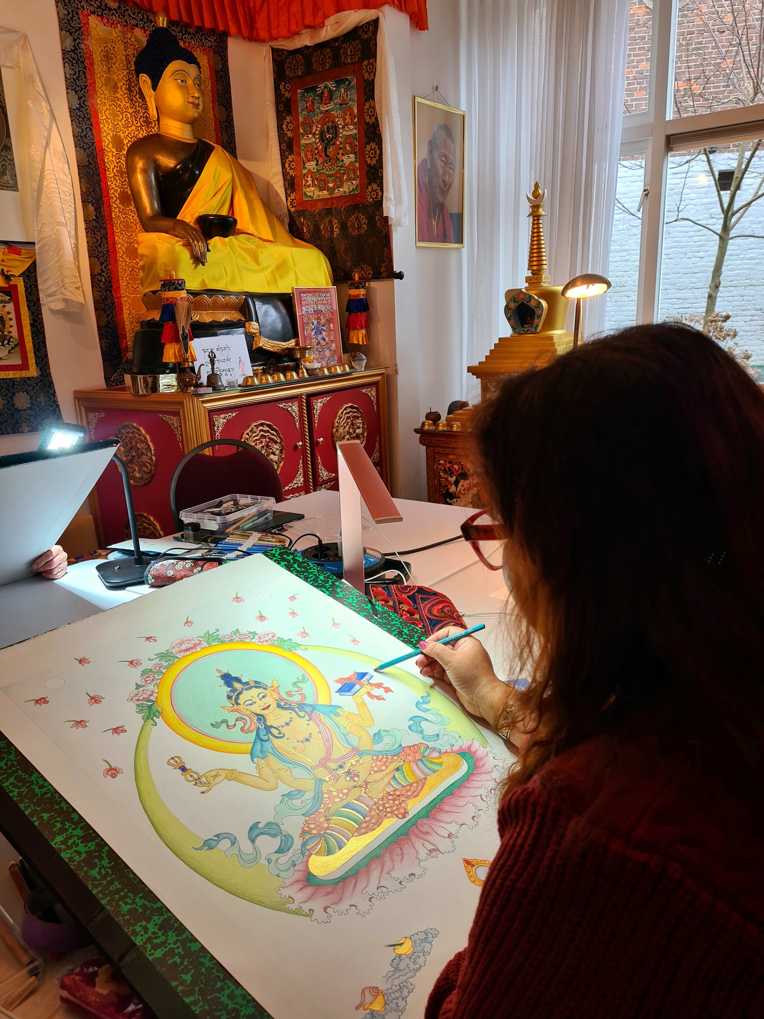 Een vrouw die een kleurrijke tanka voorstelling van een godin schildert op een tafel, met een boeddhabeeld, foto's en decoraties in de achtergrond.