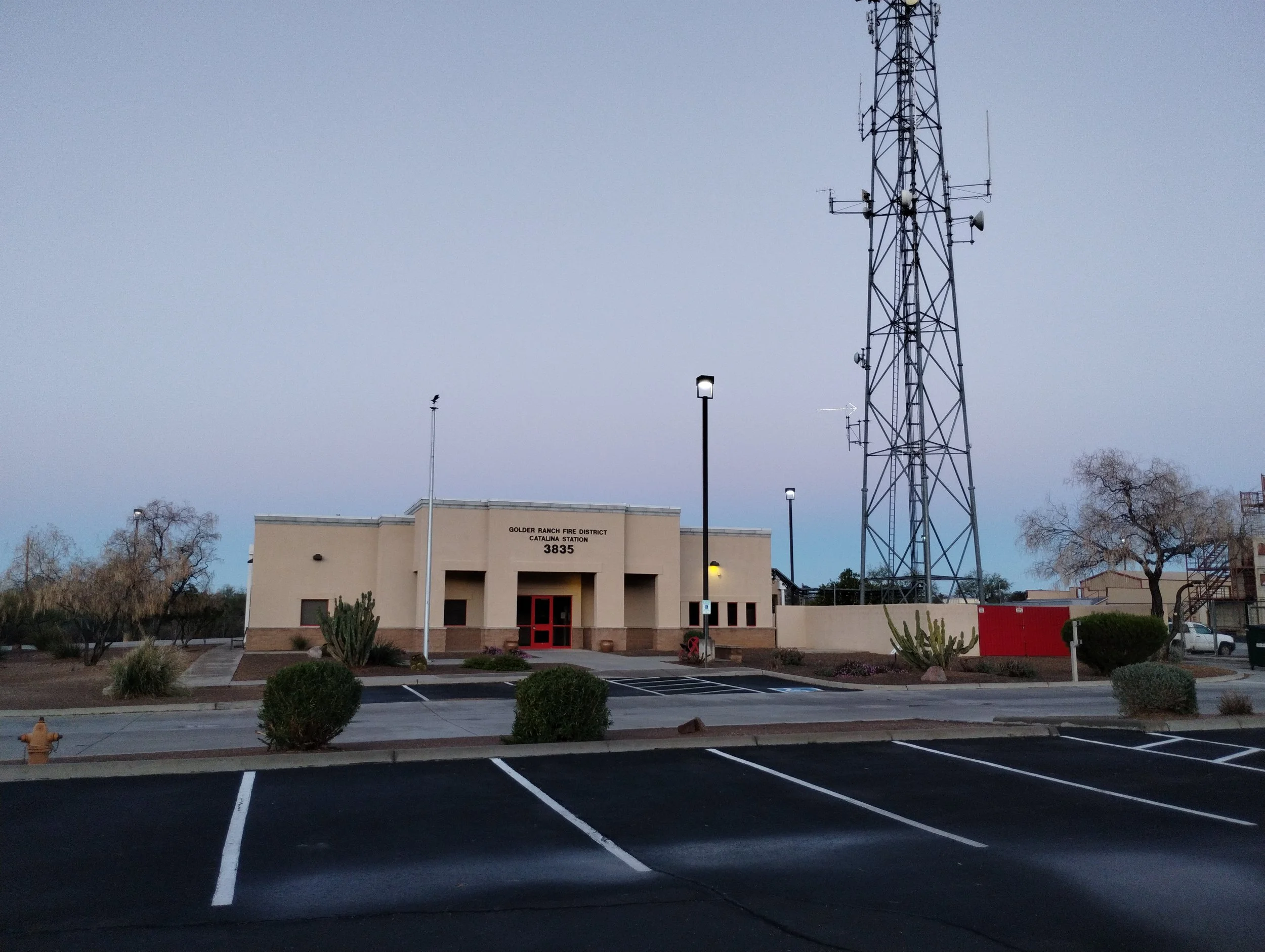 Fire station building with a sign reading "Golder Ranch Fire District Catalina Station 3835," surrounded by desert plants and parking lot, with a large radio tower and trees nearby.