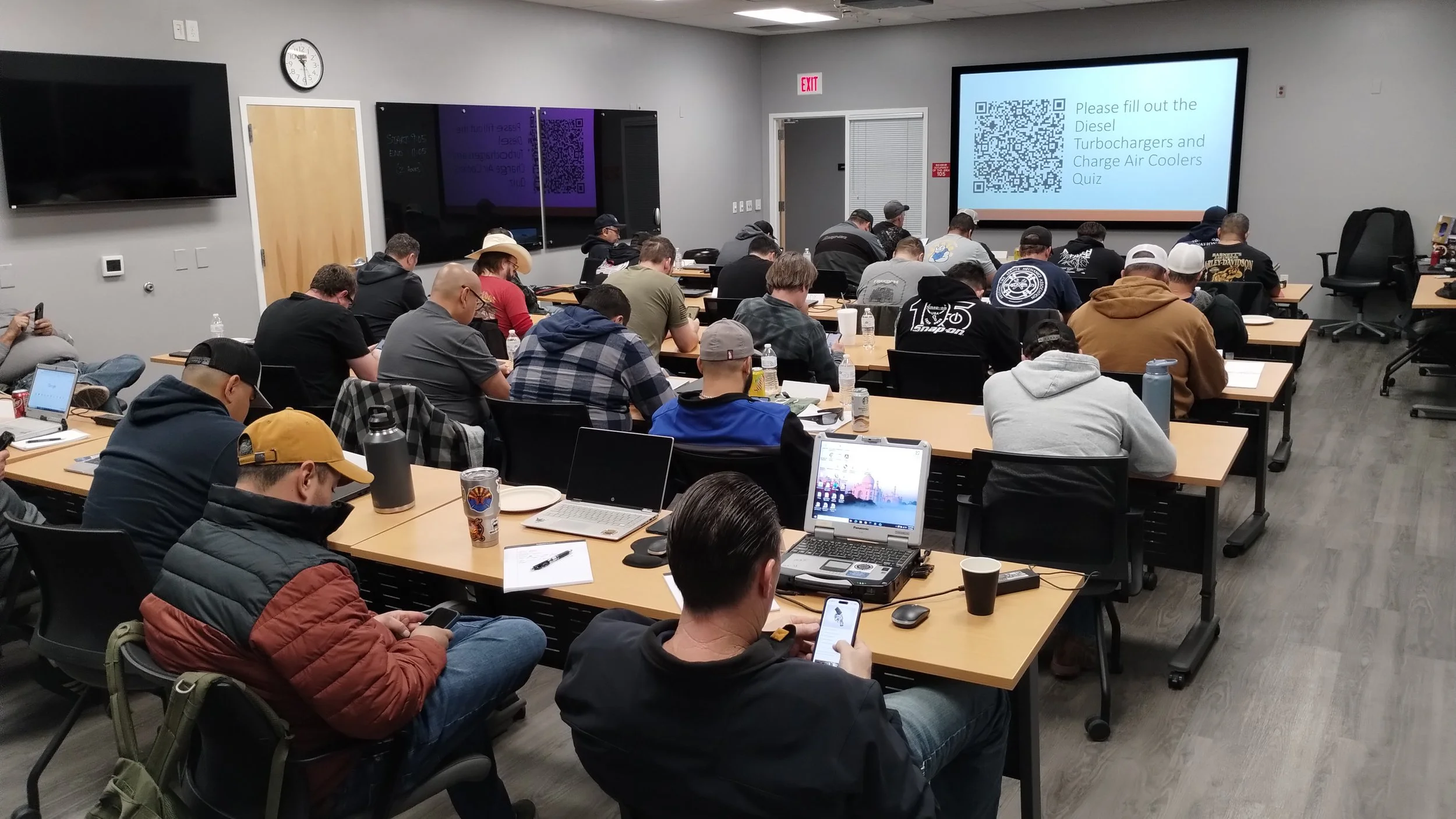 A classroom filled with EVT mechanics seated at tables, using laptops and smartphones. On the screen a QR code for a quiz.
