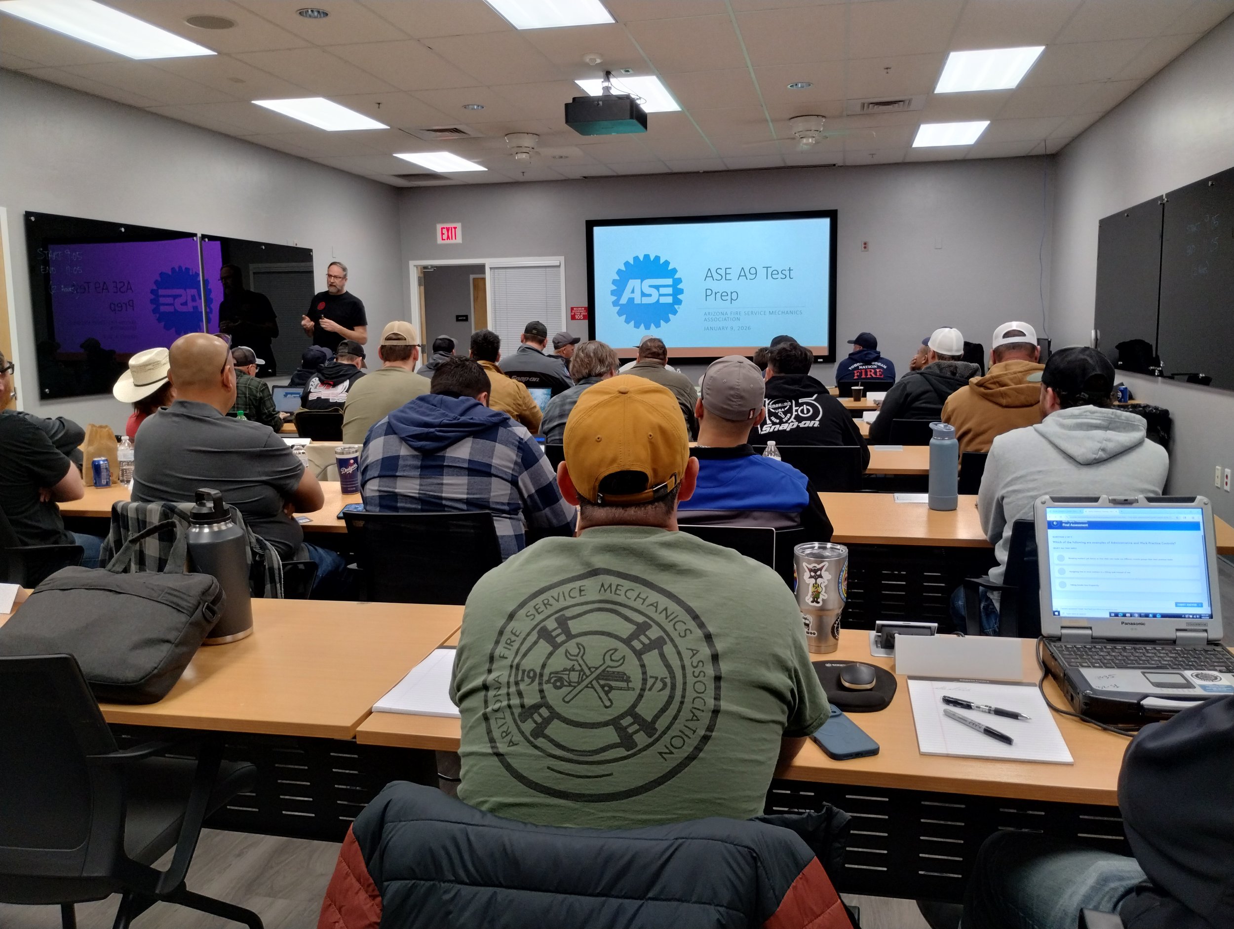 A conference room filled with technicians attending a presentation on ASE A9 Test Prep. Tony Martin stands at the front near a screen displaying the presentation title. 