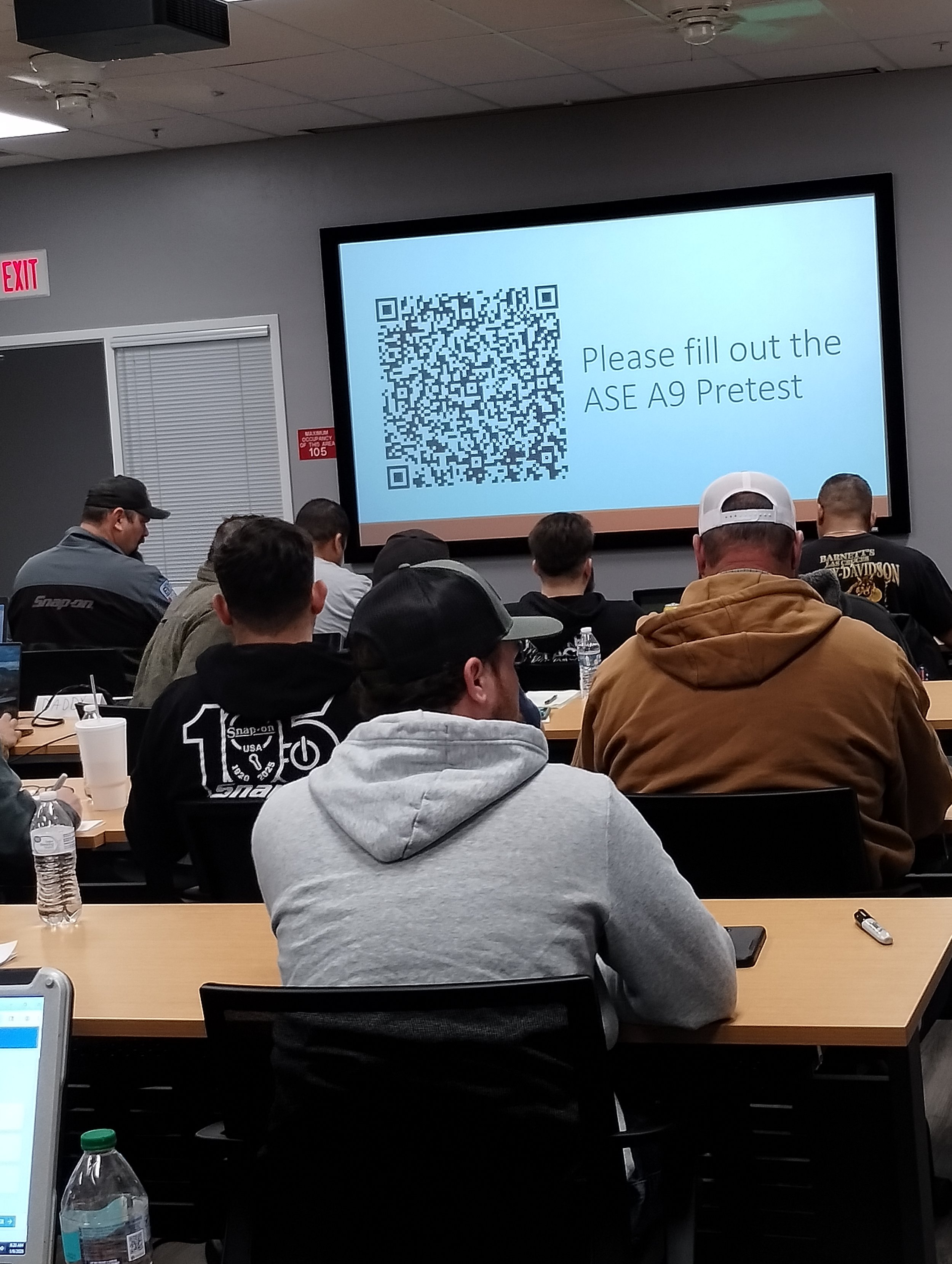A classroom of Emergency Vehicle Technicians seated at desks, facing a large screen displaying a QR code and the text 'Please fill out the ASE A9 Pretest'.