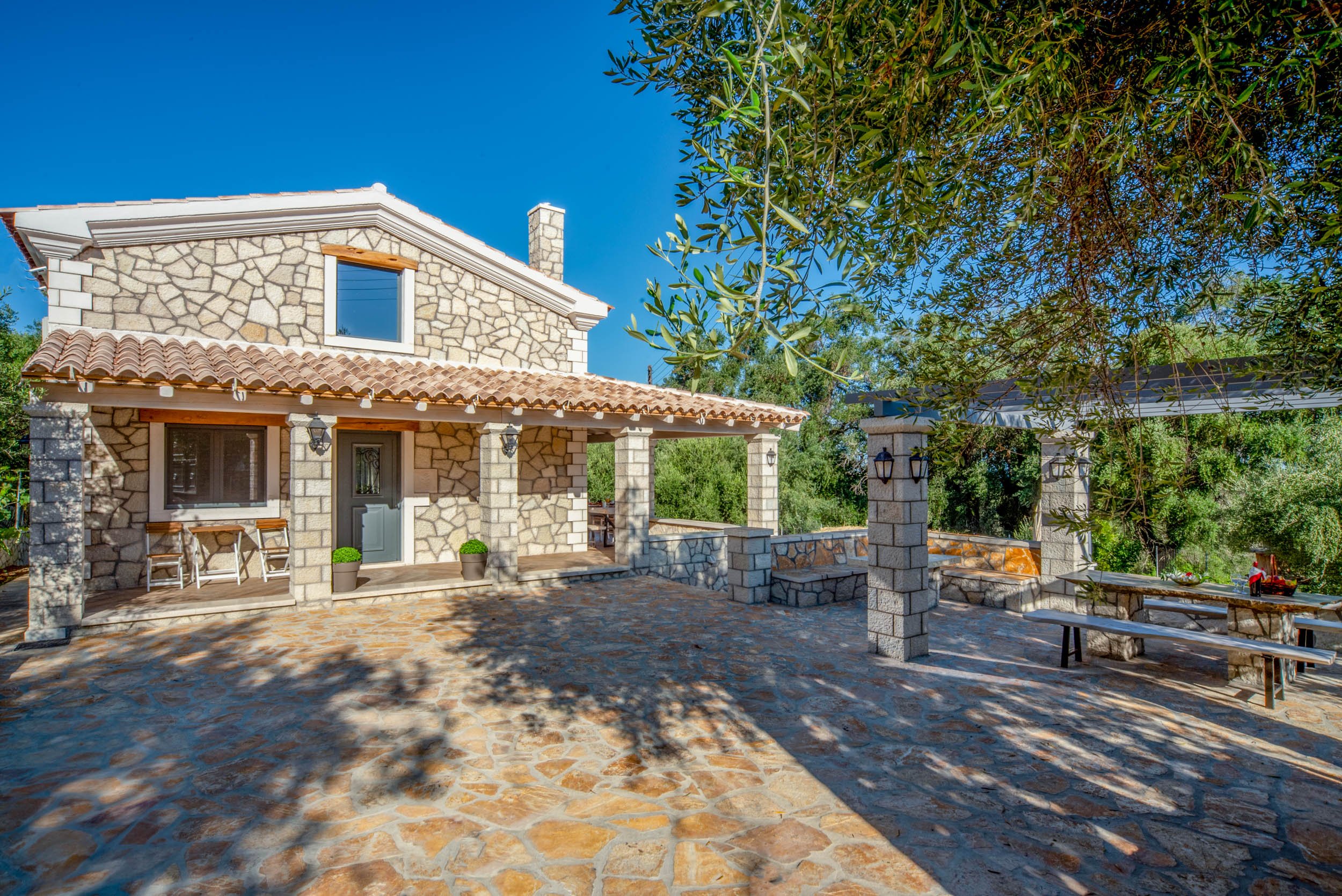 Stone house with tiled roof, porch, and outdoor seating area under a blue sky, surrounded by trees.