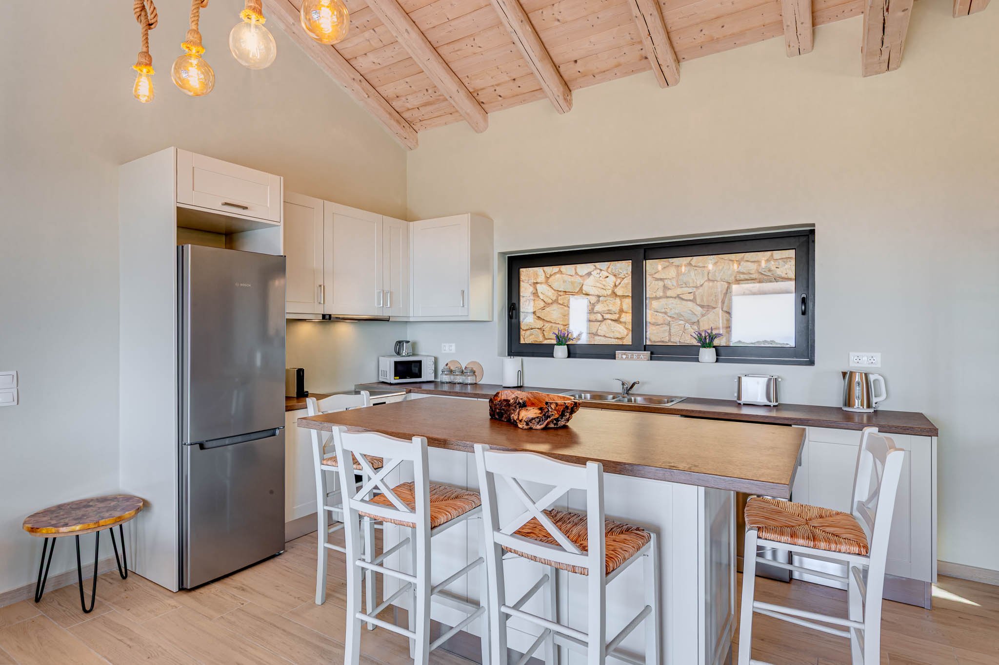 Modern kitchen with wooden ceiling, island with stools, stainless steel refrigerator, microwave, toaster, and kettle, large window, pendant lighting.