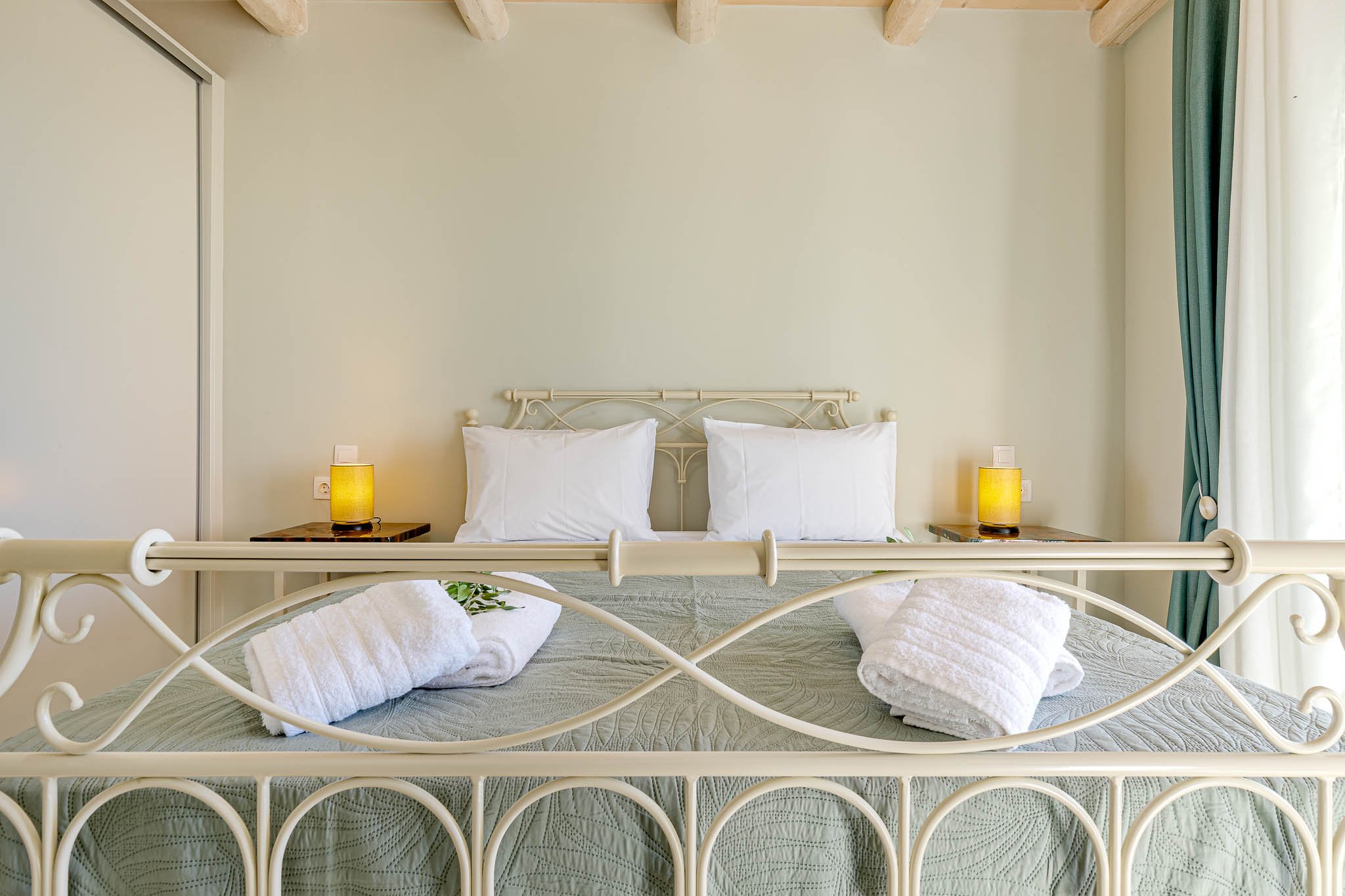 Cozy bedroom with white metal bed frame, two white pillows, folded towels on a green quilted bedspread, and yellow lamps on bedside tables. Light walls and a partially open curtain.