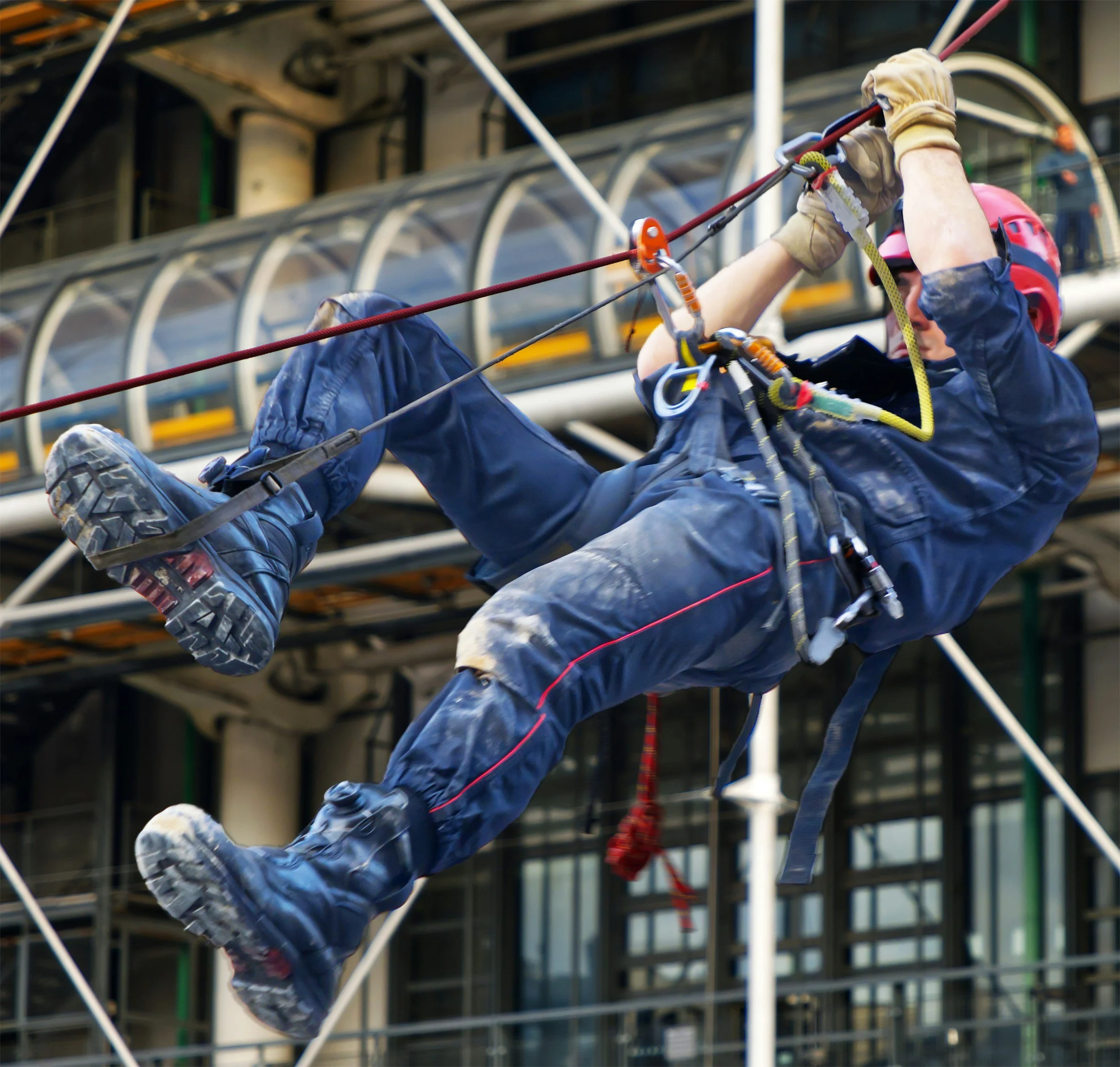 A person wearing a pink helmet, gloves, and outdoor gear is rappelling down a building while securing themselves with a rope during a rescue or maintenance operation.