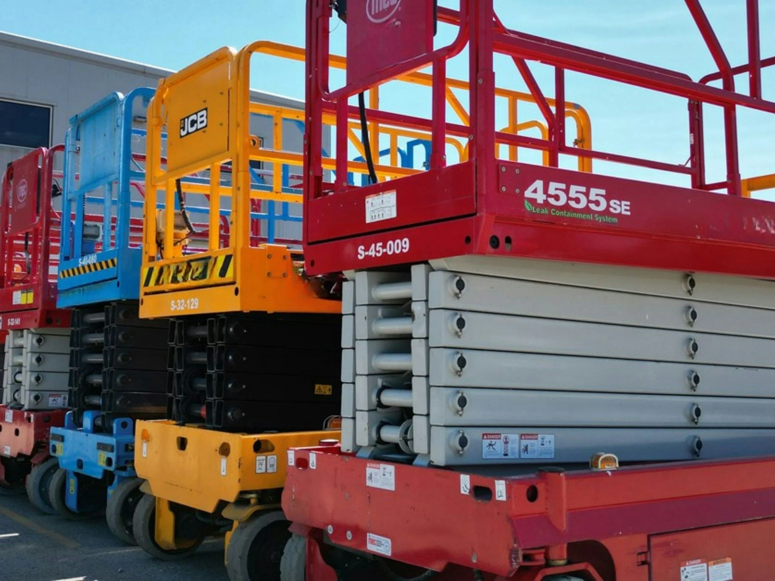 Multiple construction lifts in red, yellow, blue, and beige colors parked outdoors against a building with a clear blue sky.