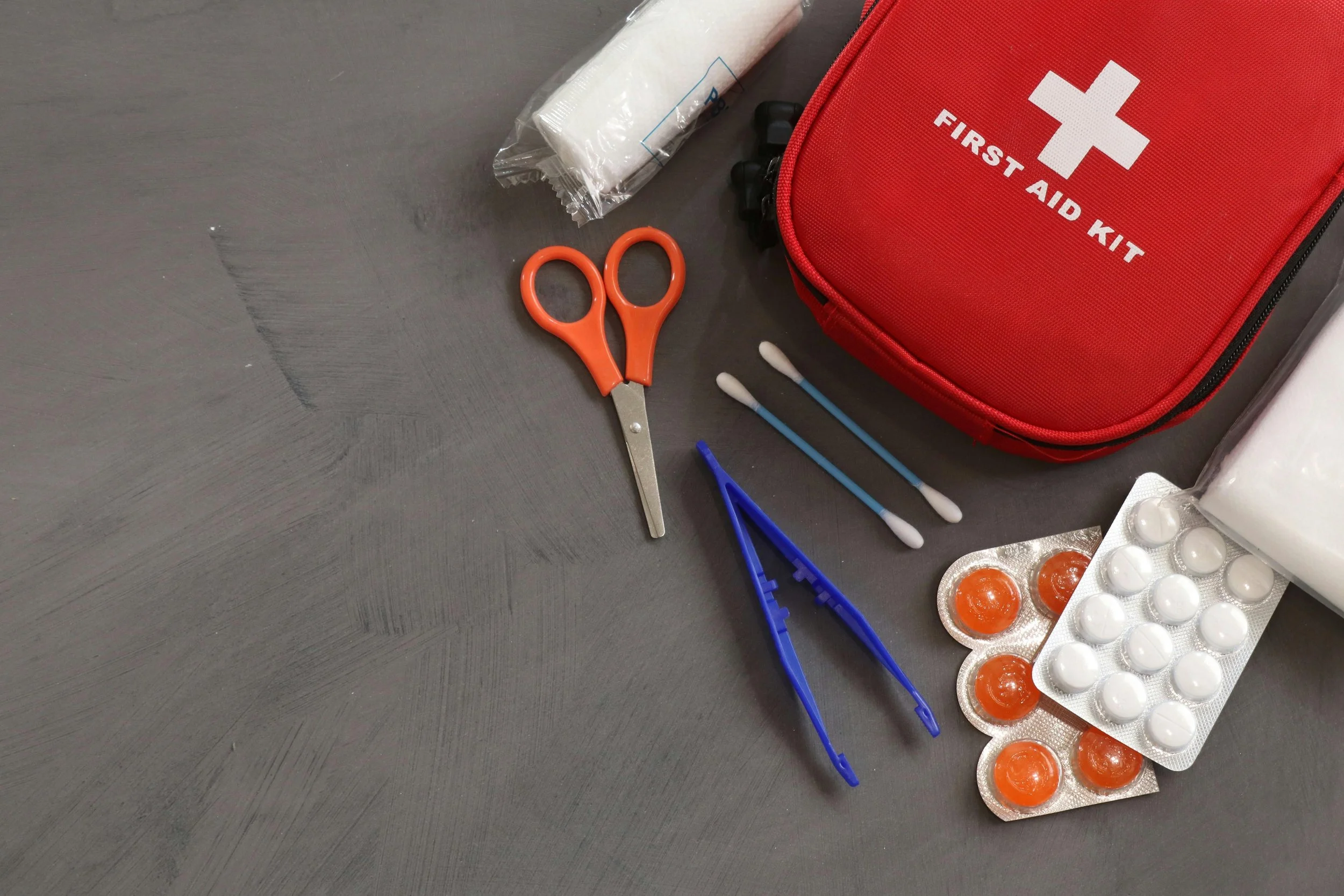 A first aid kit, a pair of orange scissors, cotton swabs, finger cots, and blister packs of pills on a dark surface.