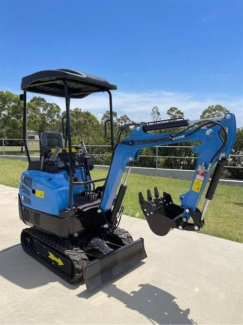 A small blue compact excavator on a concrete surface outdoors with a grassy field and trees in the background.