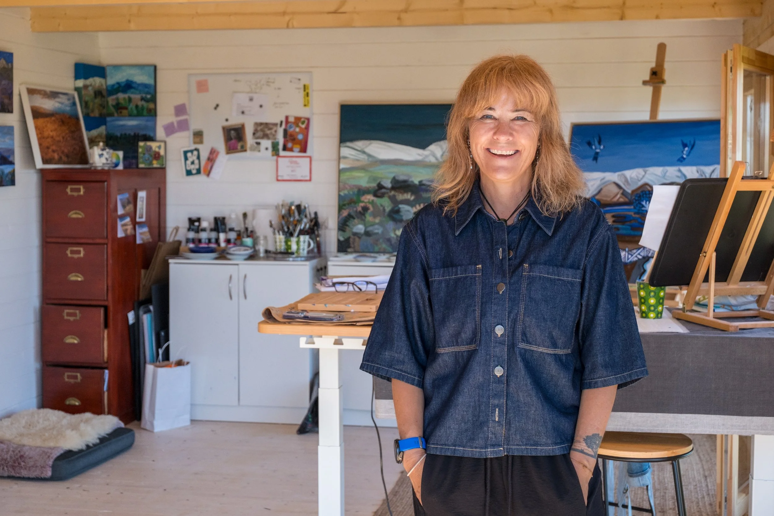 A smiling woman standing in an artist's studio, surrounded by paintings, art supplies, and easels in Central Otago.