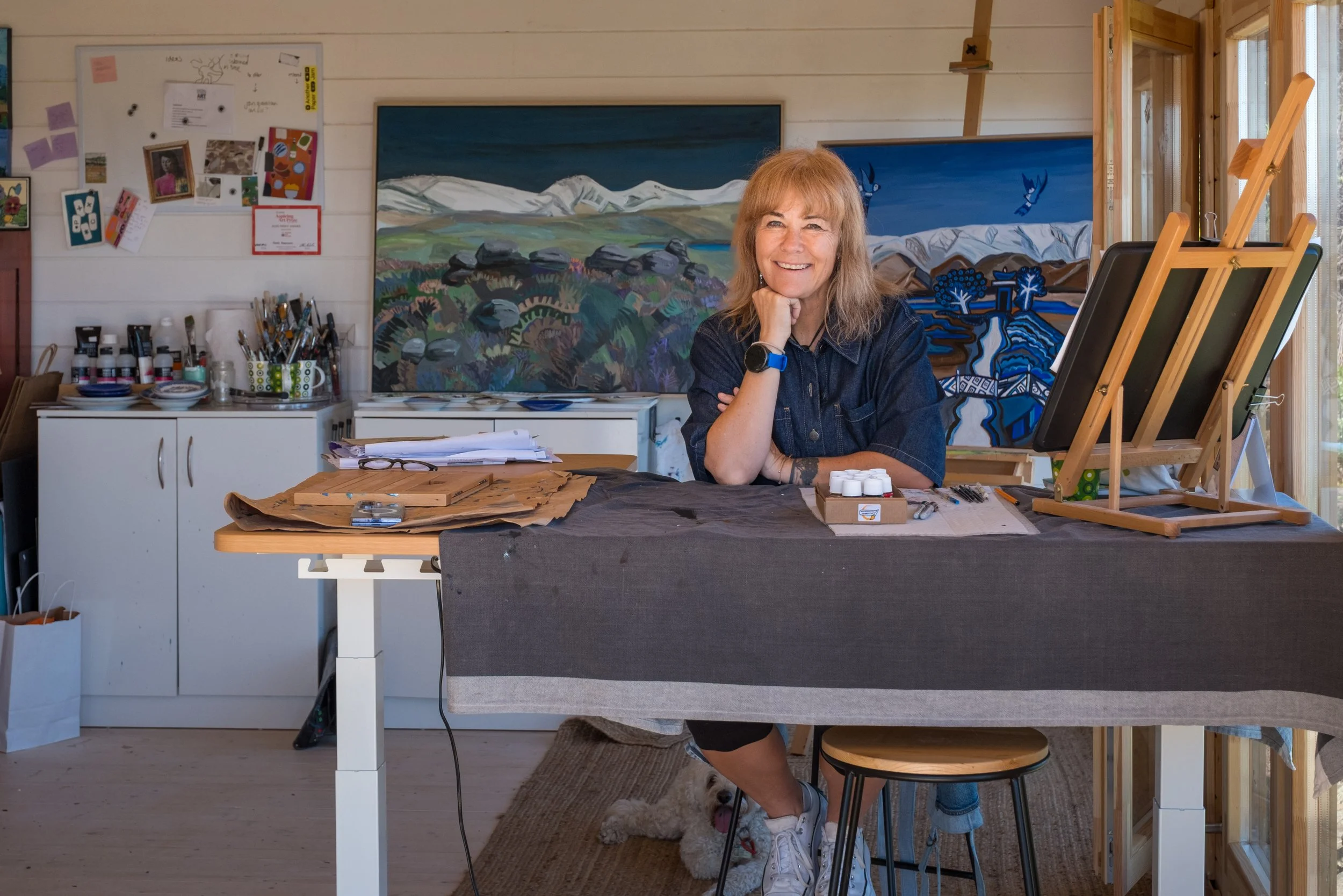 A woman sitting at an art studio table with painting supplies, smiling, with two large landscape paintings behind her, and a dog lying on the floor underneath the table.