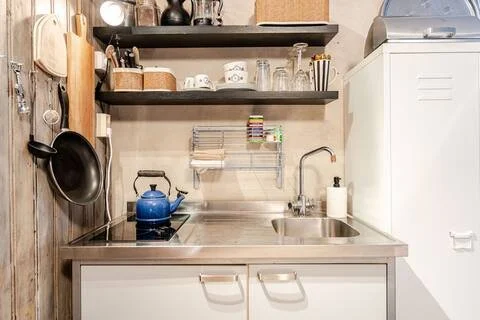 Een keuken met een gootsteen, een fornuis met een blauwe ketel, en wandplanken met verschillende items zoals pannen en glazen.