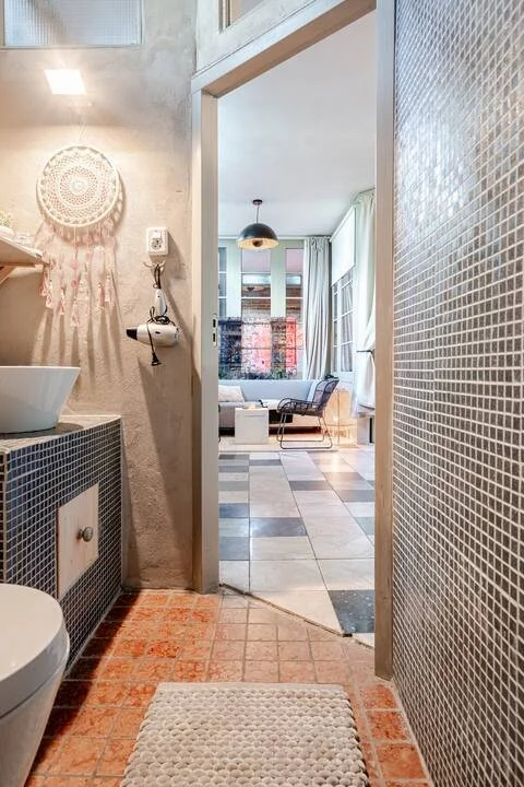 Kleine badkamer met wastafel, toilet en decoratieve muurhanger, opening naar een lichte woonkamer met zitgedeelte, grote ramen en uitzicht.