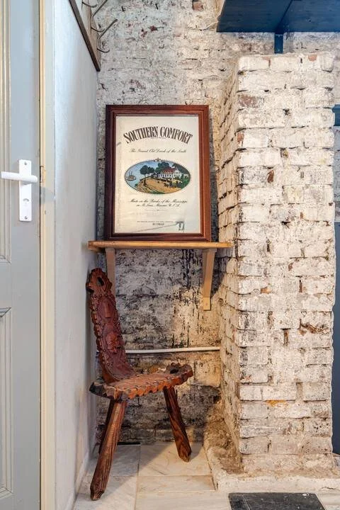 Een zwart bord met de tekst Southerny Comfort en een afbeelding van een huis en een meer, geplaatst op een houten plank tegen een bakstenen muur in een interieur.