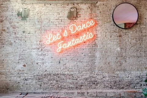 Een gemetselde muur met een neonlichttekst die zegt 'Let's Dance Fantastic' en een ronde spiegel rechtsboven.