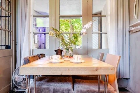 Een lichte eetkamertafel met thee- of koffiekommen en een vaas met bloemen in een zonnige kamer met grote ramen.
