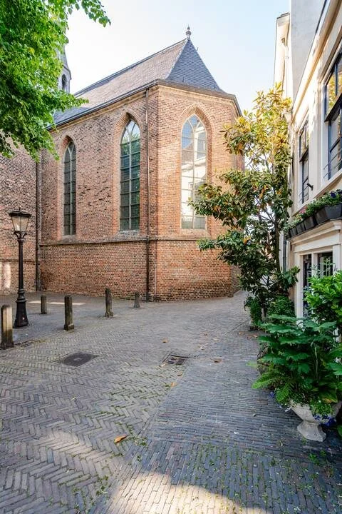 Een open, smal straatje met een kerk-achtige stenen gebouw met grote raampartijen en een spitse dak, omgeven door bomen en traditionele stadsgebouwen.