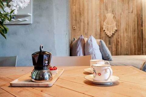 Een koffiezetapparaat en twee kopjes met schotels op een houten eettafel in een gezellige, moderne woonkamer met een houten muur en kussens op een bank.