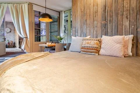 Een gezellige slaapkamer met een grote bed, open ramen, en een houten wand, met een kleine eettafel en planten.