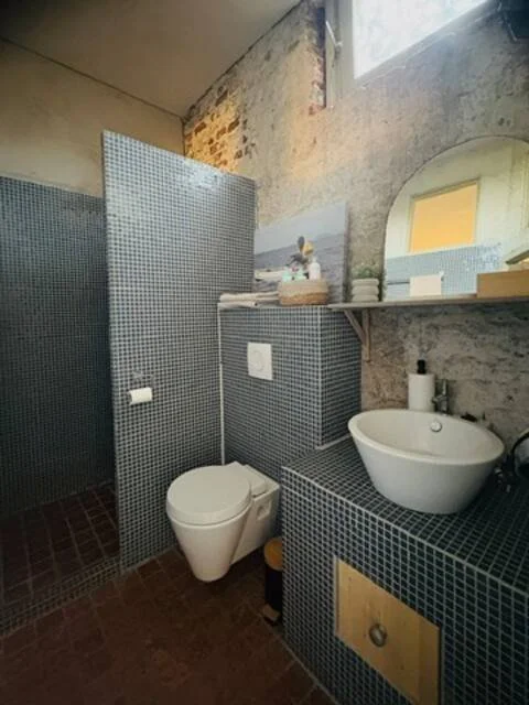 Een kleine badkamer met muur- en vloerbedekking van kleine blauwe tegels, een toilet, een wastafel met een ronde witte wasbak en een spiegel, een plank met handdoeken en toiletartikelen, en een raam met een gele gordijn.