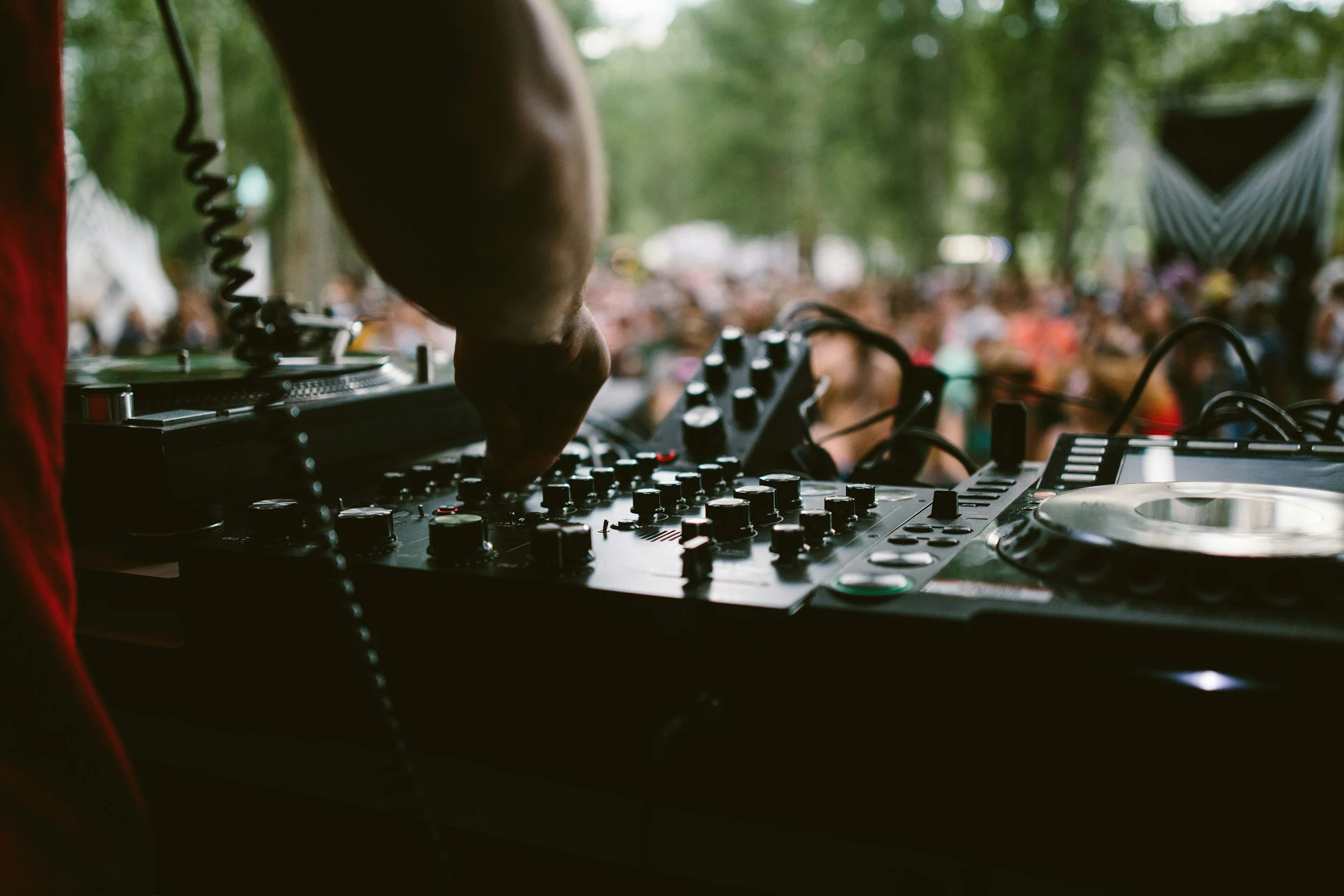 Close-up of a DJ's hand on a mixer at an outdoor music festival, with a blurred crowd and trees in the background.