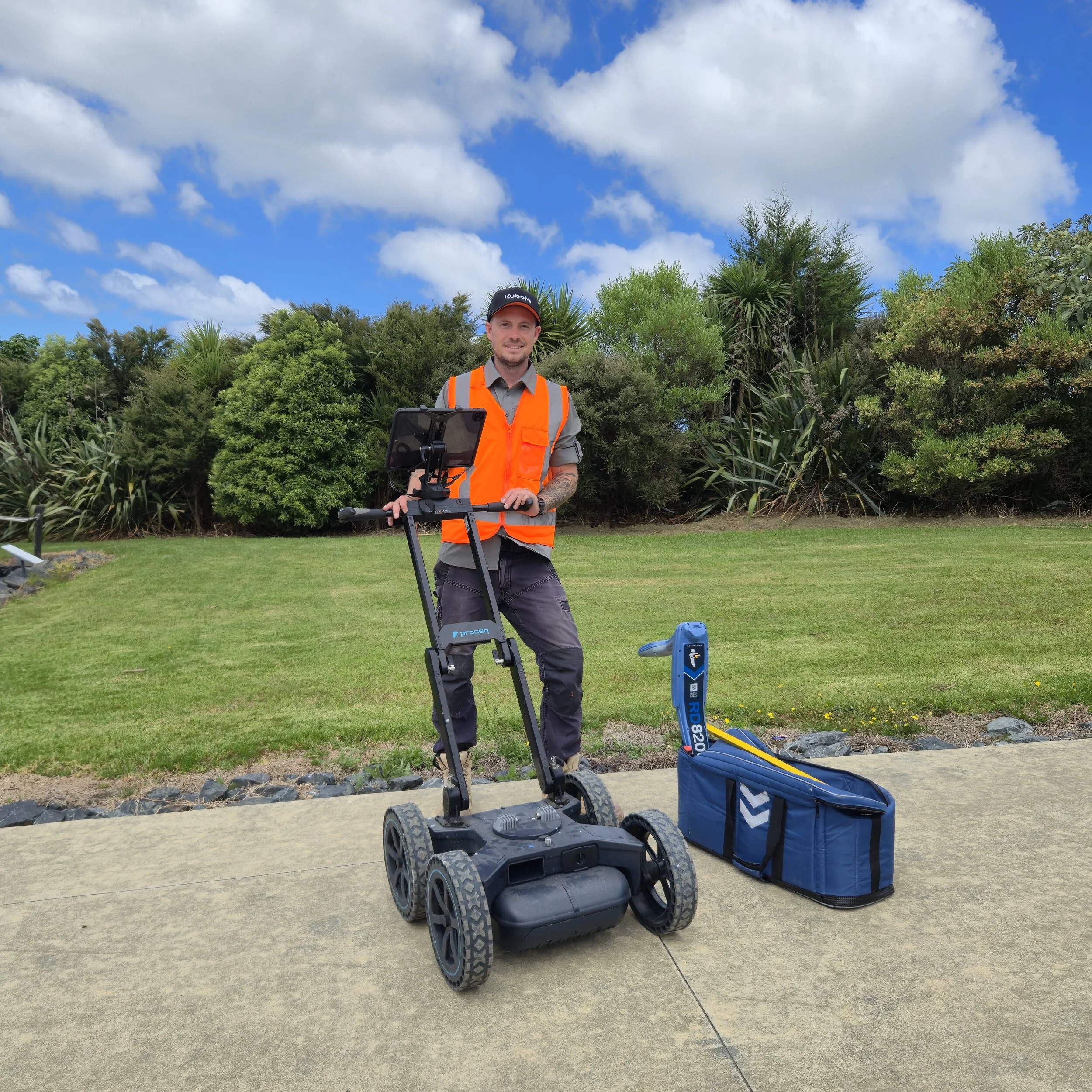 A man in an orange safety vest and gray shirt stands outdoors with a ground-penetrating radar device, surrounded by green trees and a partly cloudy sky.