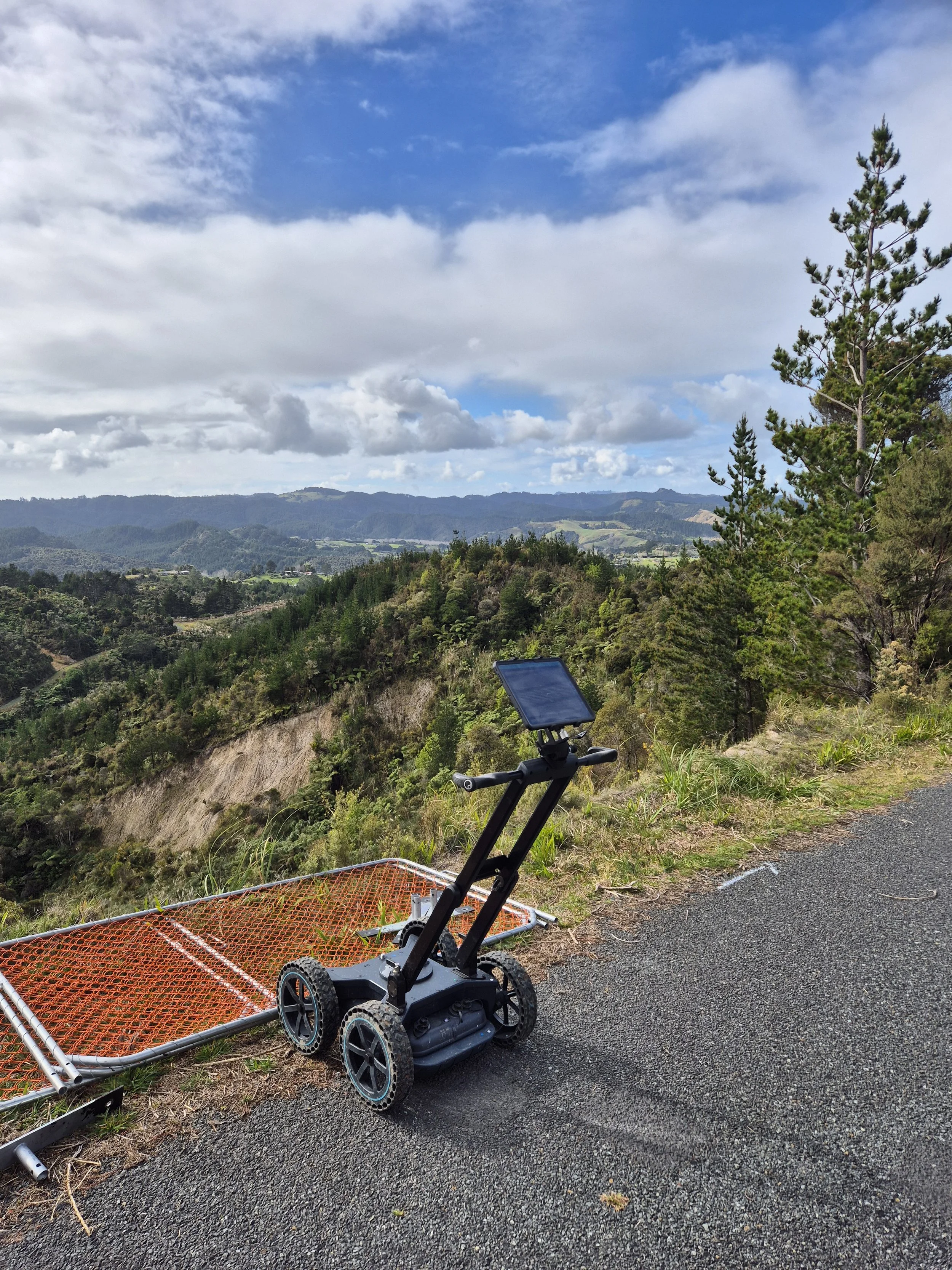 A robotic device with a touchscreen, four wheels, and a handlebar, parked next to a fallen orange safety barrier on the side of a mountain road, overlooking a lush, green valley under a partly cloudy sky.