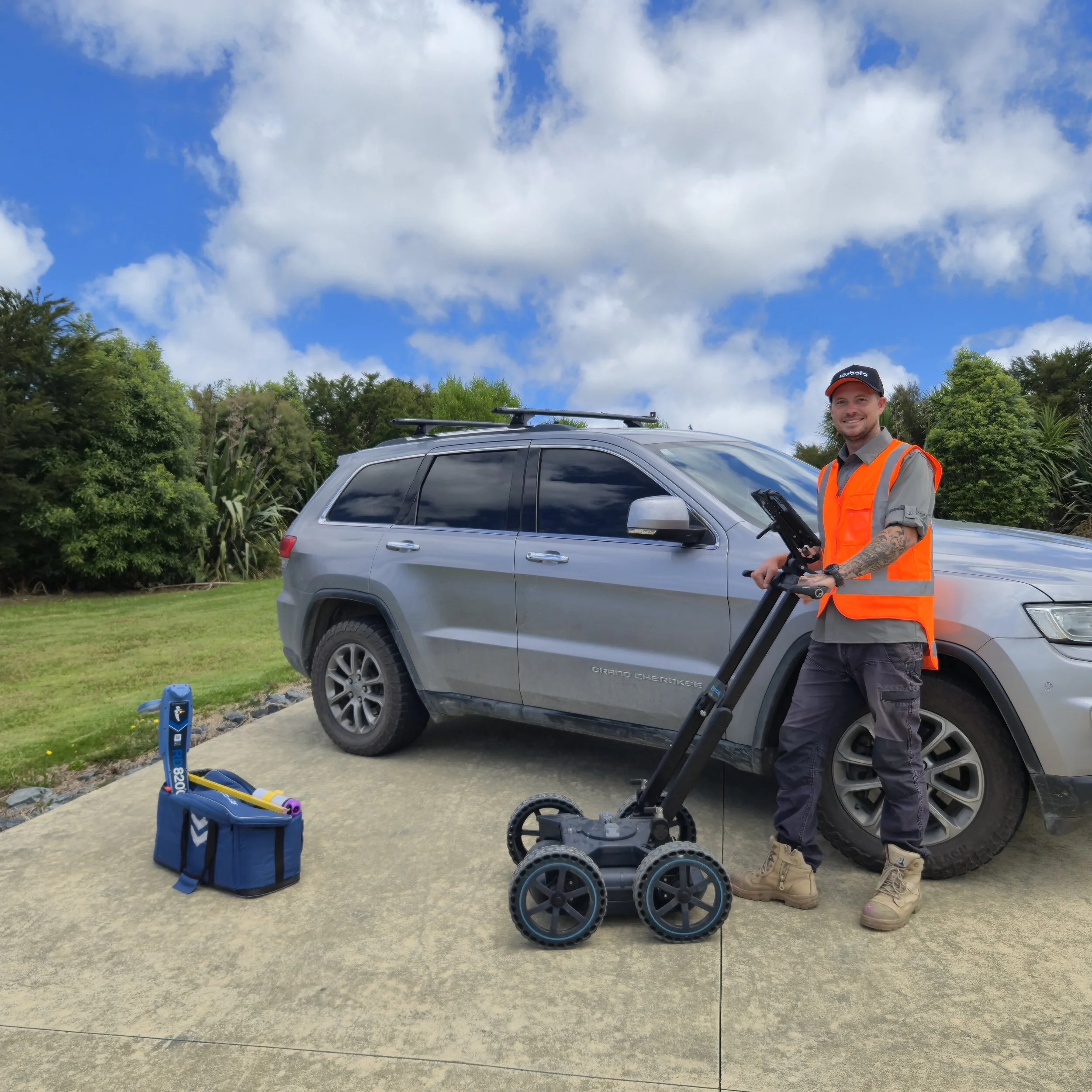 A man wearing an orange safety vest and a cap stands next to a gray Jeep car on a driveway, holding a piece of equipment. There is a robotic device on wheels and a blue toolbox with tools on the ground nearby. The background has green trees and a partly cloudy blue sky.