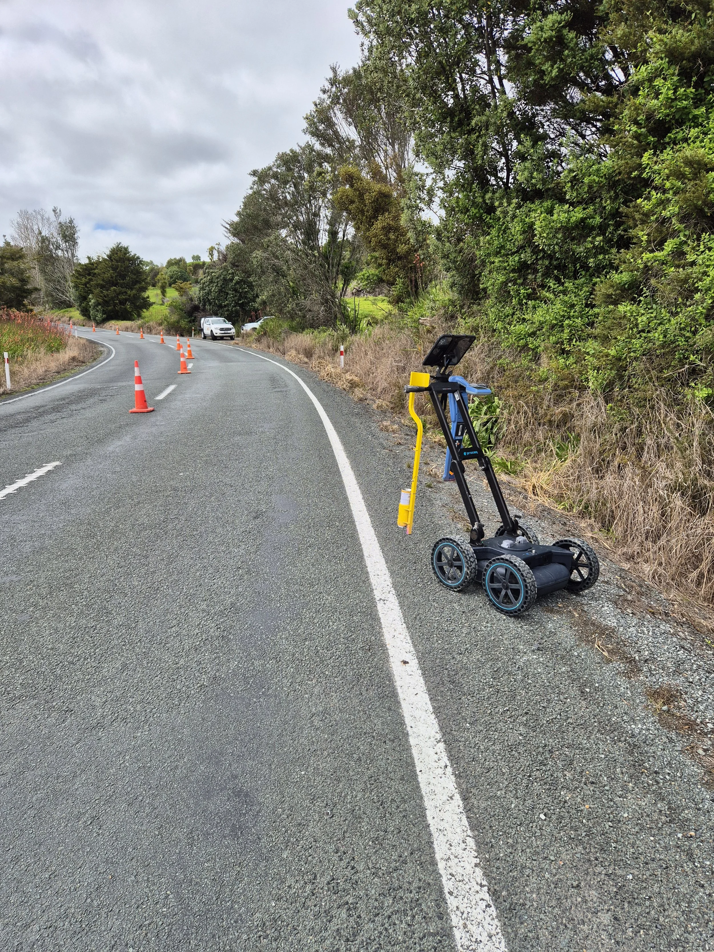 A portable speed radar sign on the side of a winding road, with orange traffic cones directing traffic, and lush green trees lining the roadside under a cloudy sky.