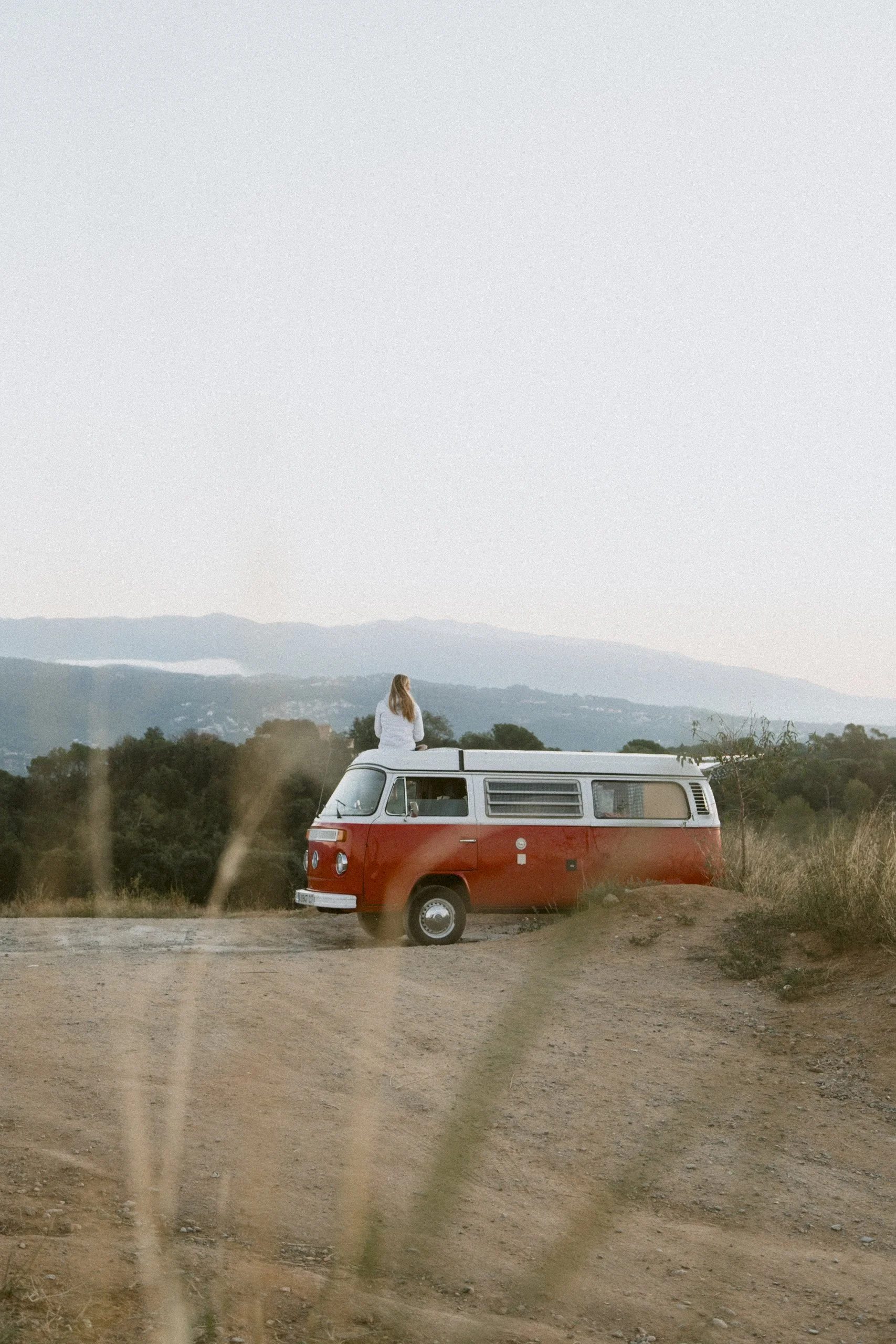 A woman with long blonde hair sitting on top of a vintage red and white camper van on a dirt hill, overlooking a landscape of trees and mountains during dusk.