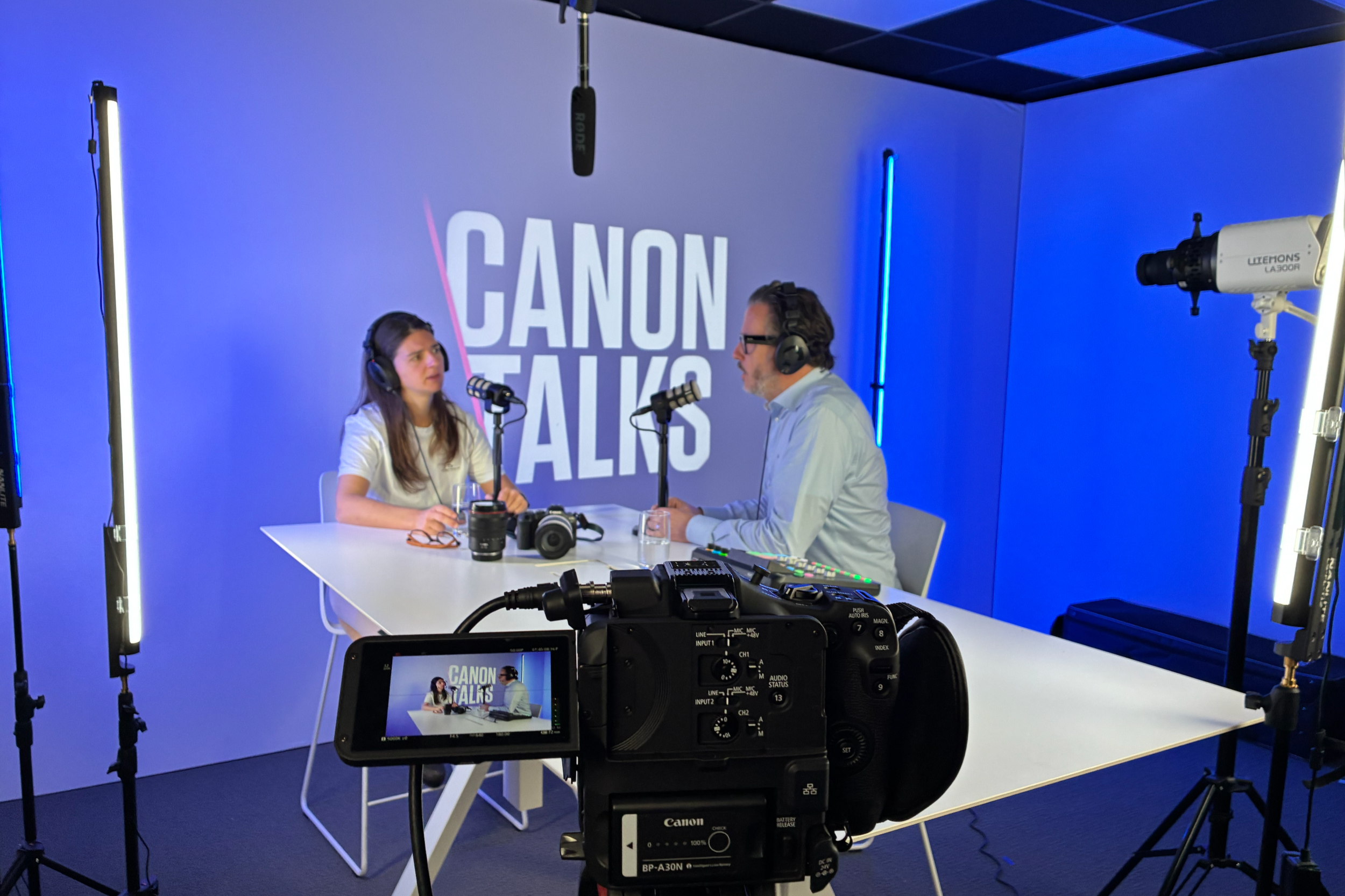Two people recording a podcast or interview in a studio with a blue background that says 'Canon Talks.' The woman on the left has long brown hair, glasses, and headphones, and the man on the right has curly hair, glasses, and headphones. There are pr