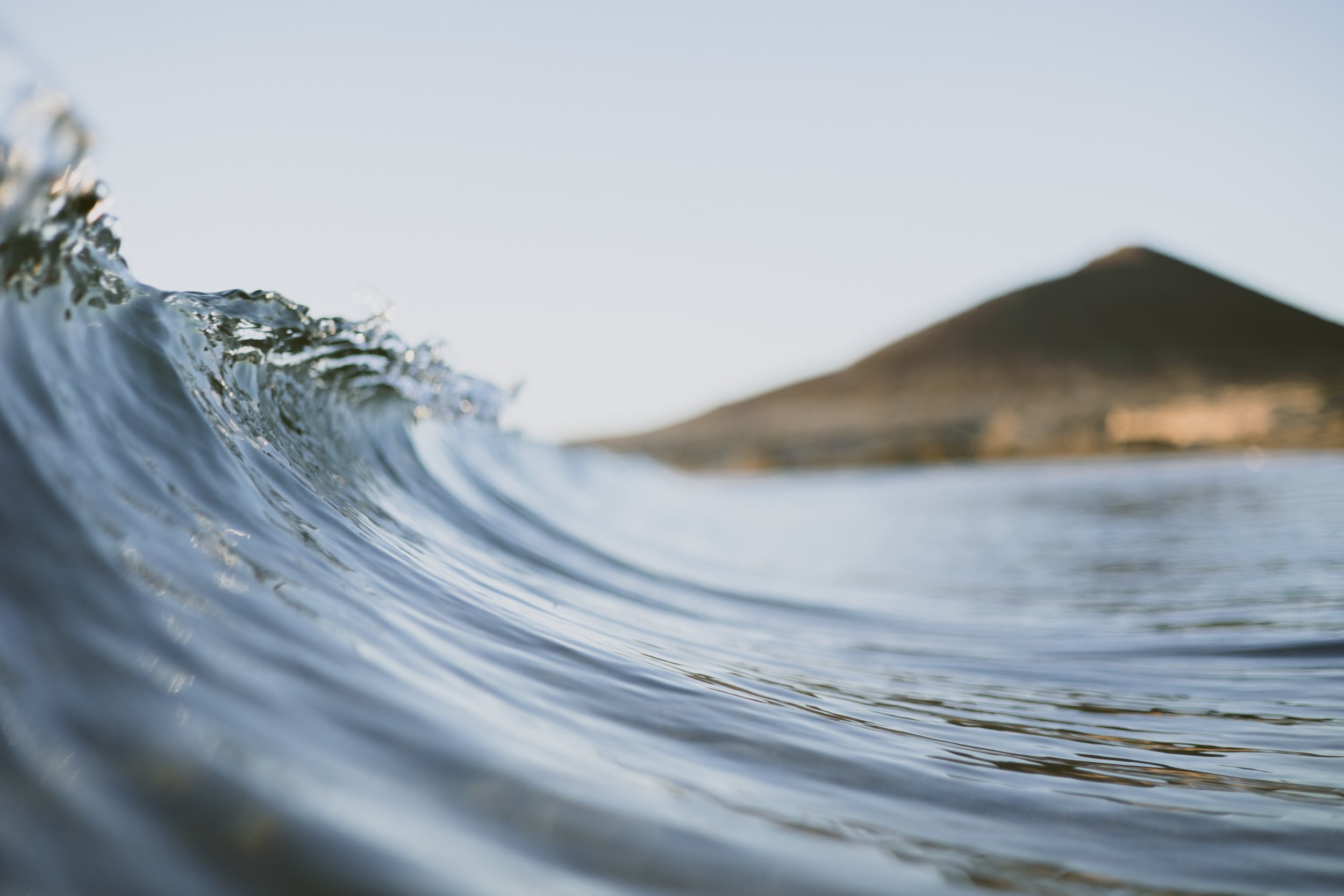 Close-up of a small ocean wave with a landmass or mountain in the background under clear sky.