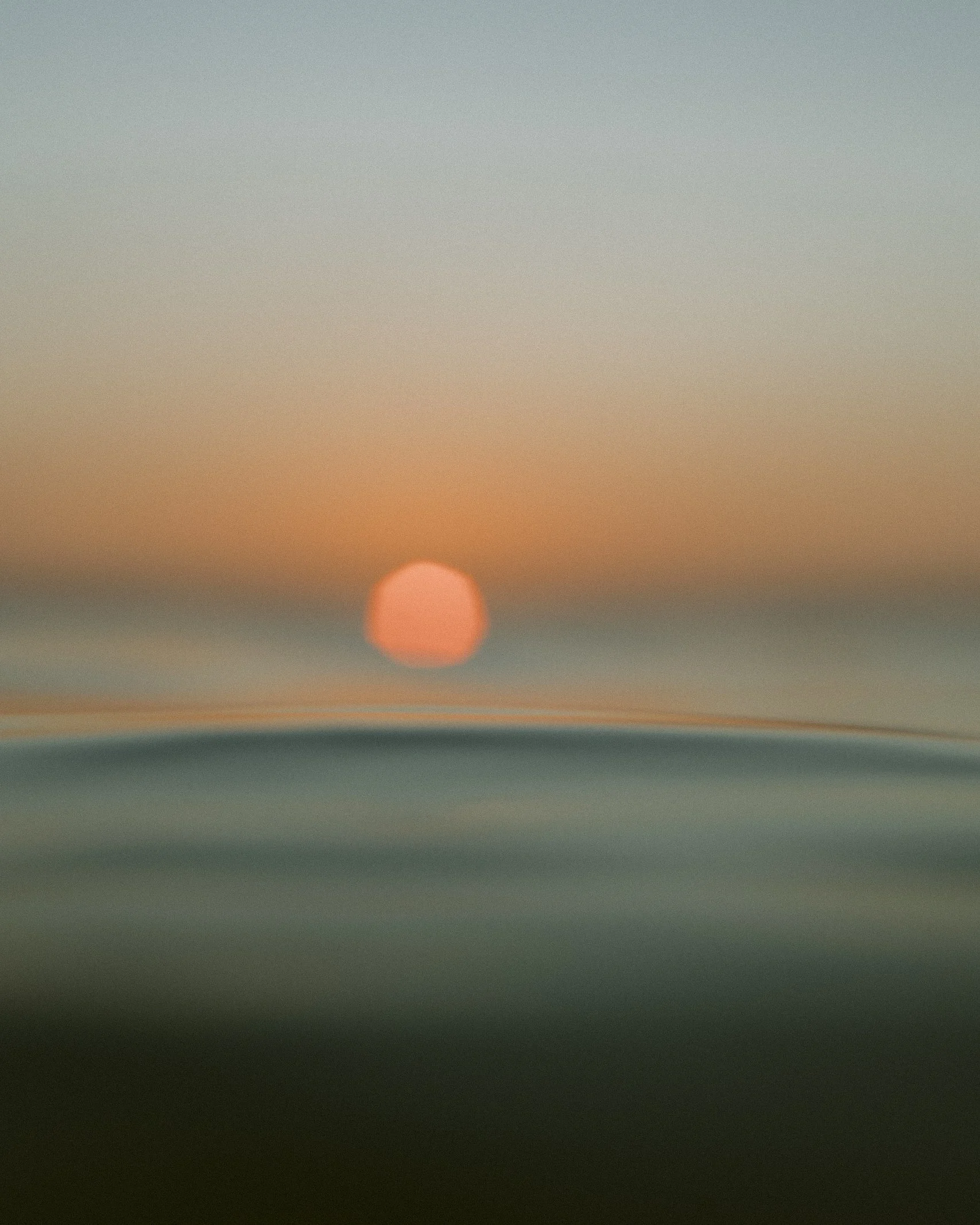 Blurred image of a sunset over the ocean with the sun near the horizon and waves in the foreground.