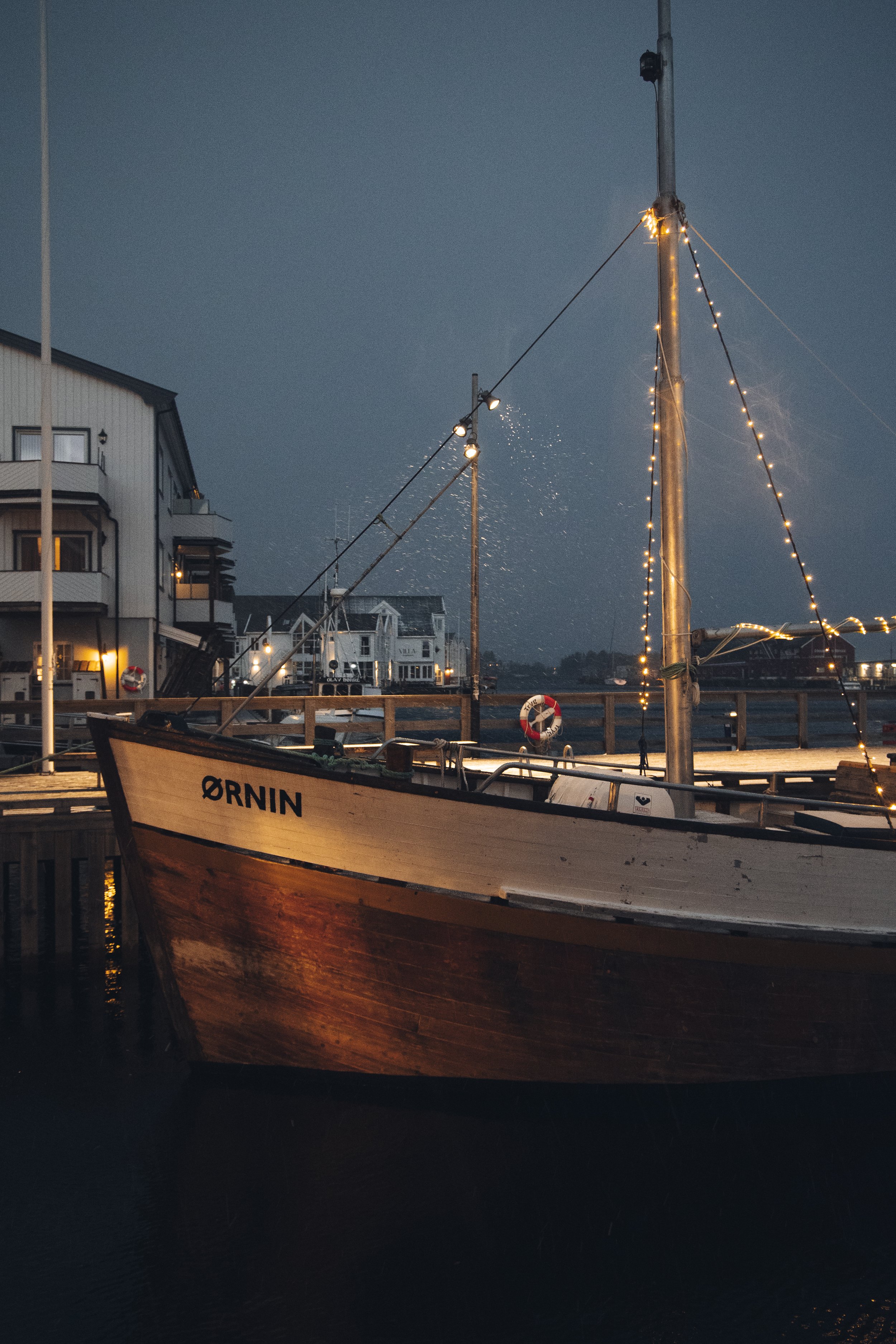 A wooden sailboat named Ørninn docked at a pier at dusk, with decorative string lights on the mast and nearby buildings illuminated.