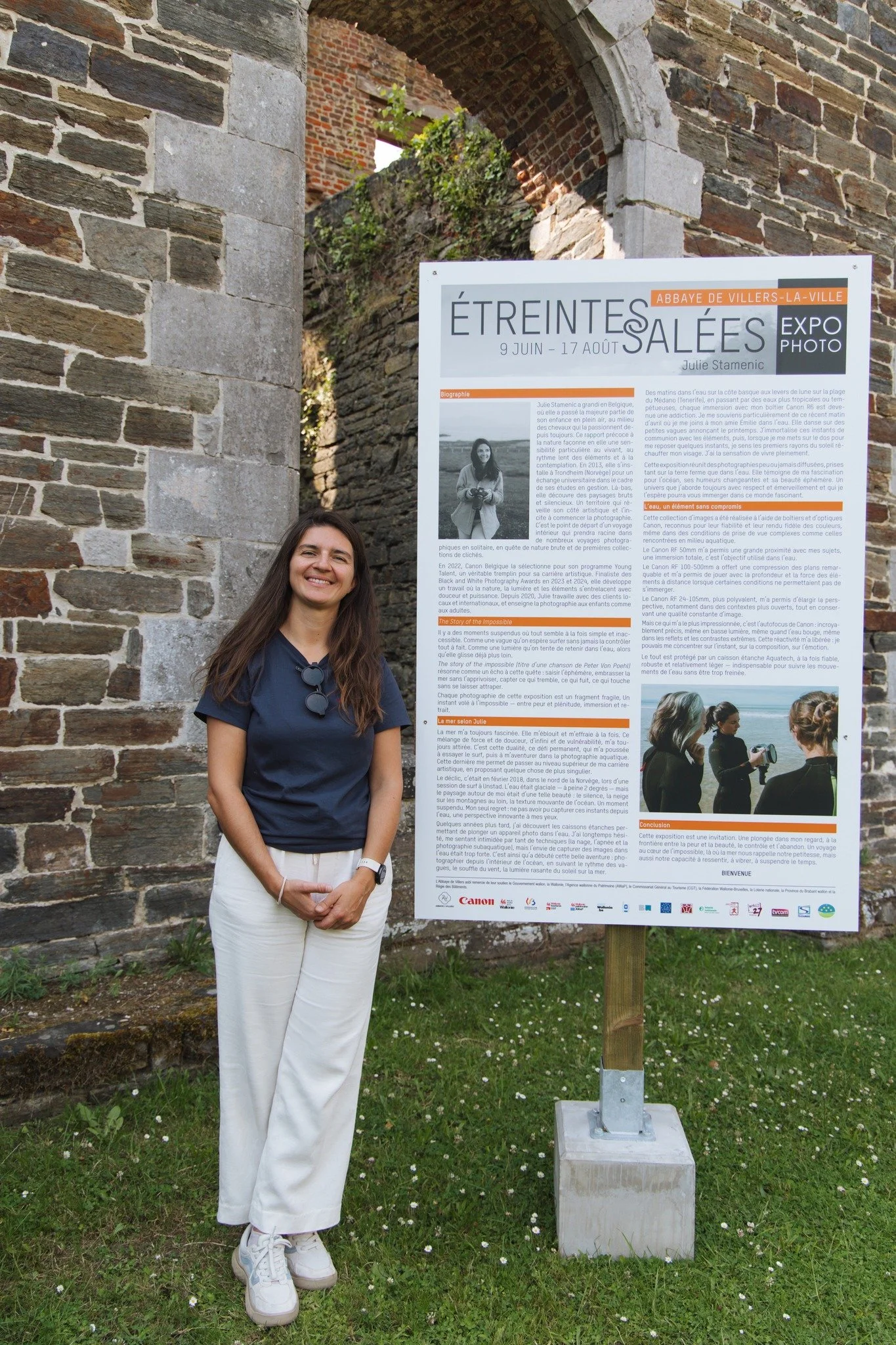 A smiling woman in a navy blue t-shirt and white pants standing outside near a large informational sign at an outdoor event or exhibition. The sign is titled 'Étreintes Salées' and includes text, photos, and logos. The backdrop features part of a his