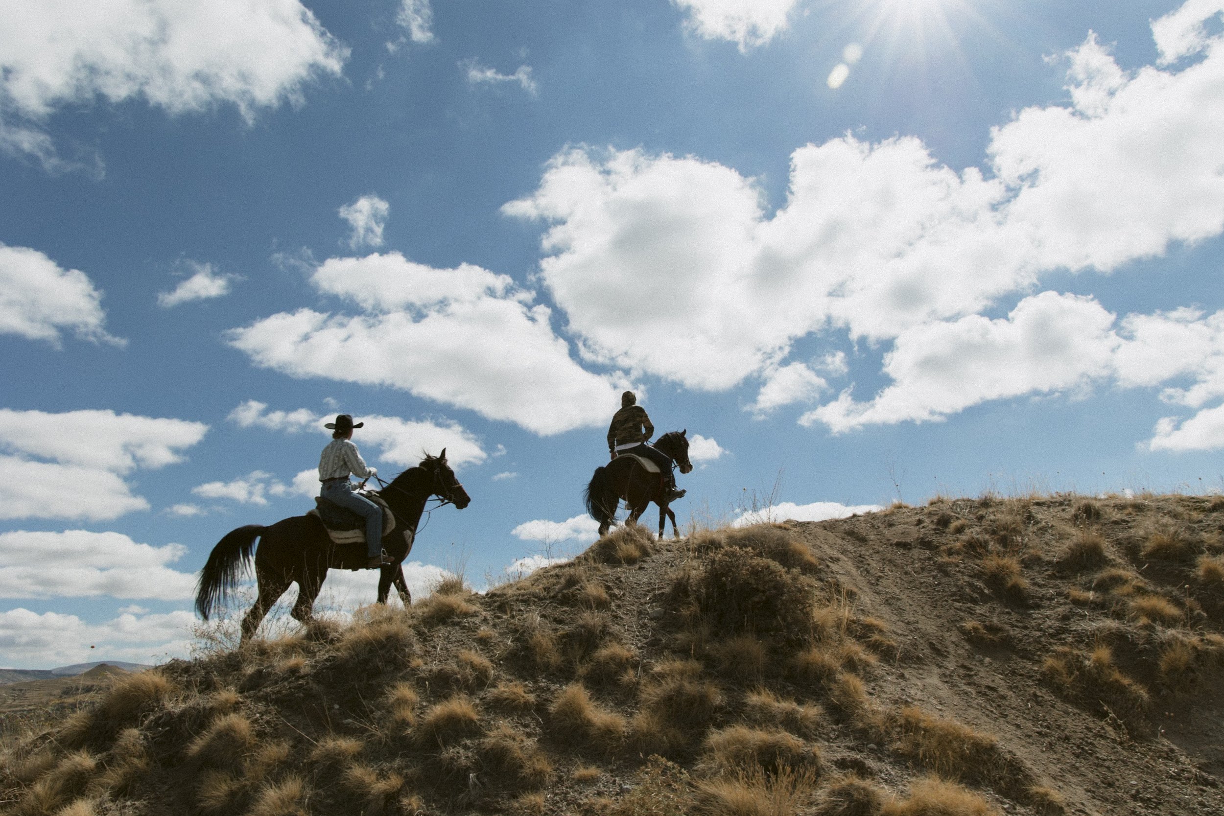Two people riding horses up a hillside with dry grass, under a partly cloudy sky with sunlight.