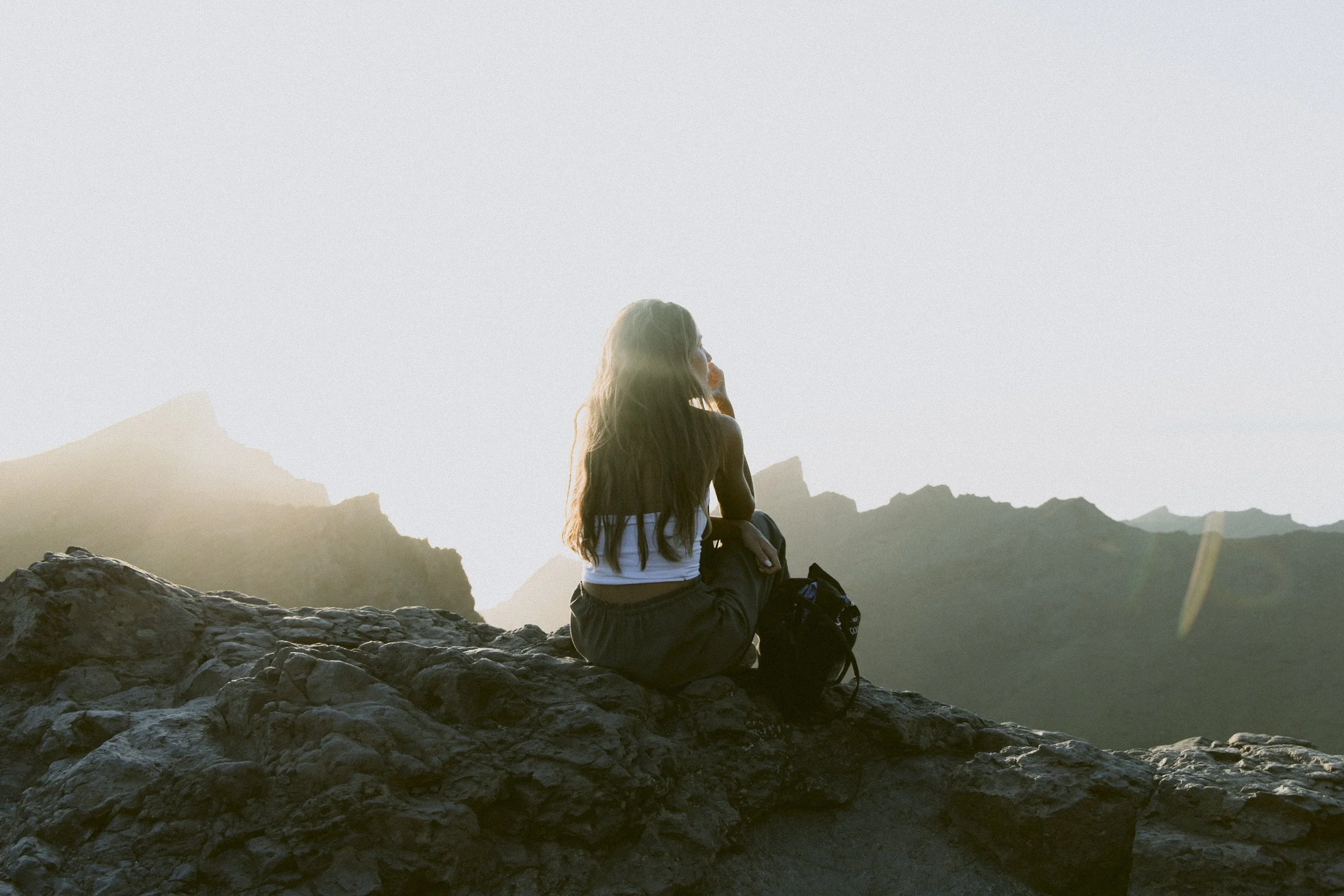 A woman with long hair sitting on a rocky ledge, gazing at mountains at sunset or sunrise.