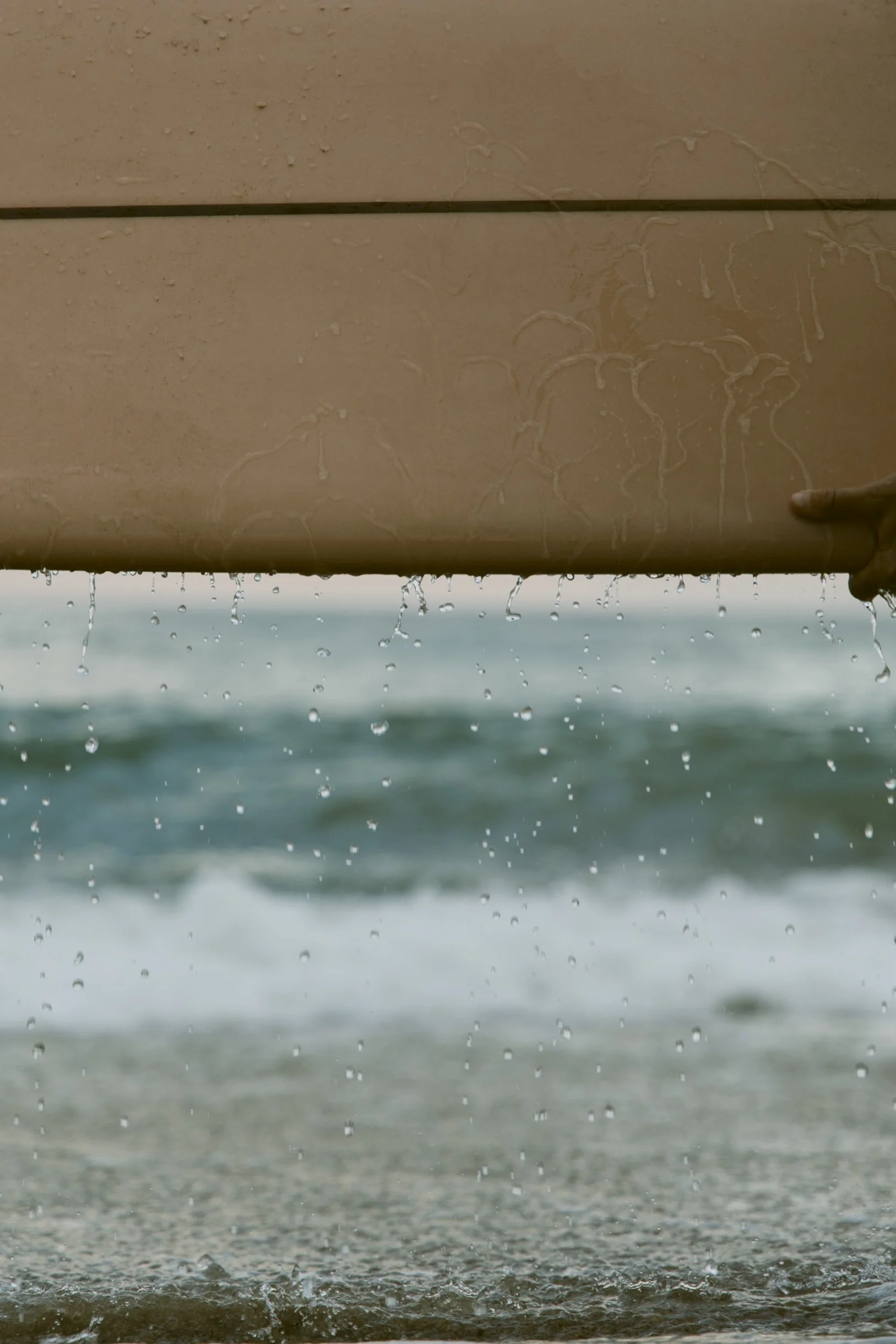 Close-up of a wooden surfboard with water droplets falling from it, with the ocean and waves in the background.
