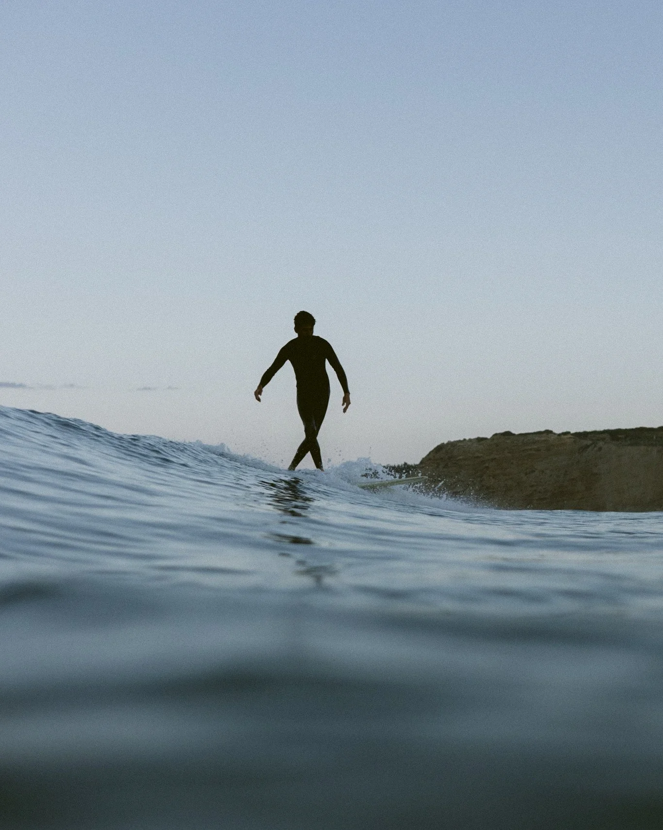 Silhouetted person in black wetsuit standing on a surfboard in the water near a rocky shoreline, with a clear sky overhead.