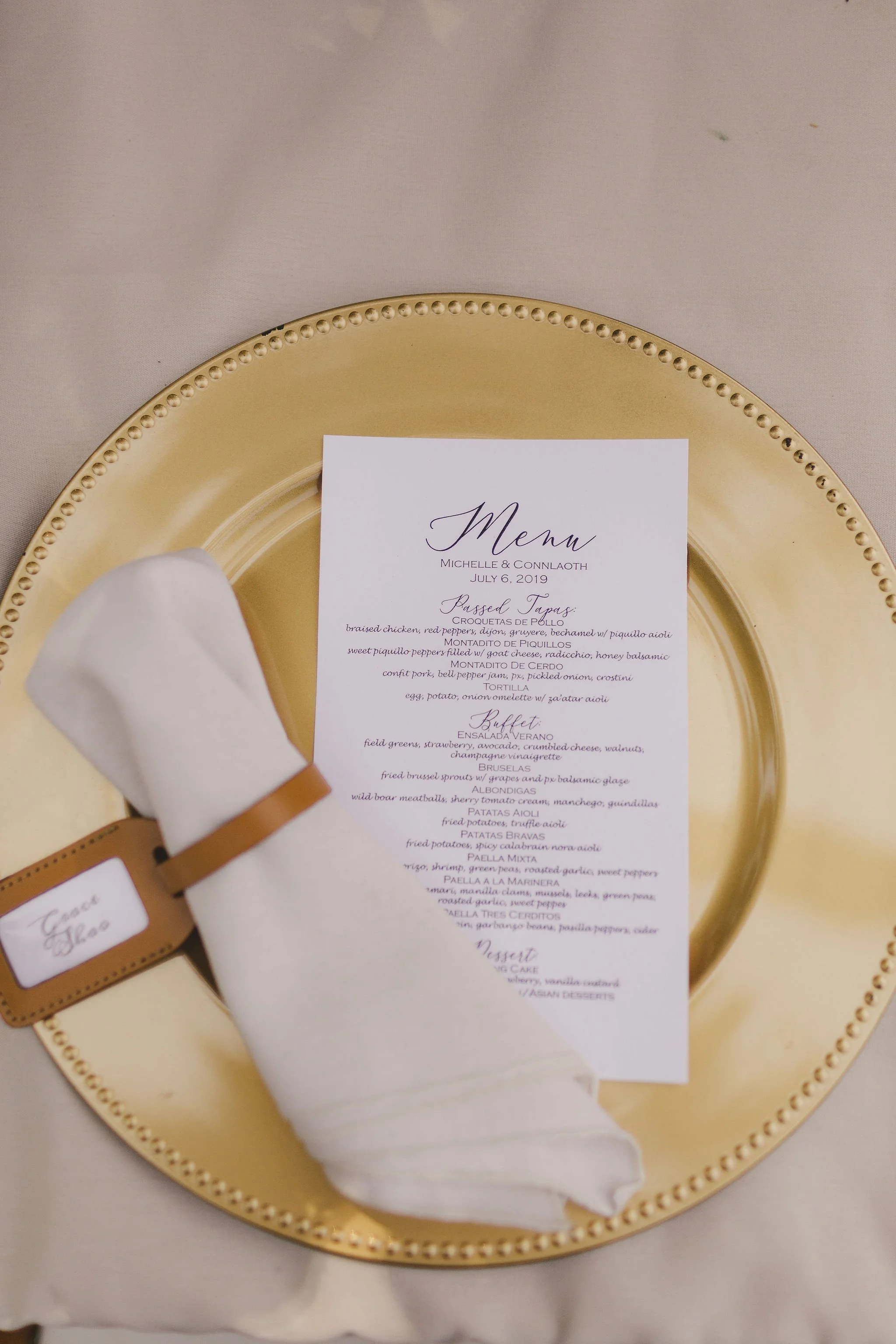 Gold charger plate with a wedding menu and a cloth napkin with a place card on a beige tablecloth.