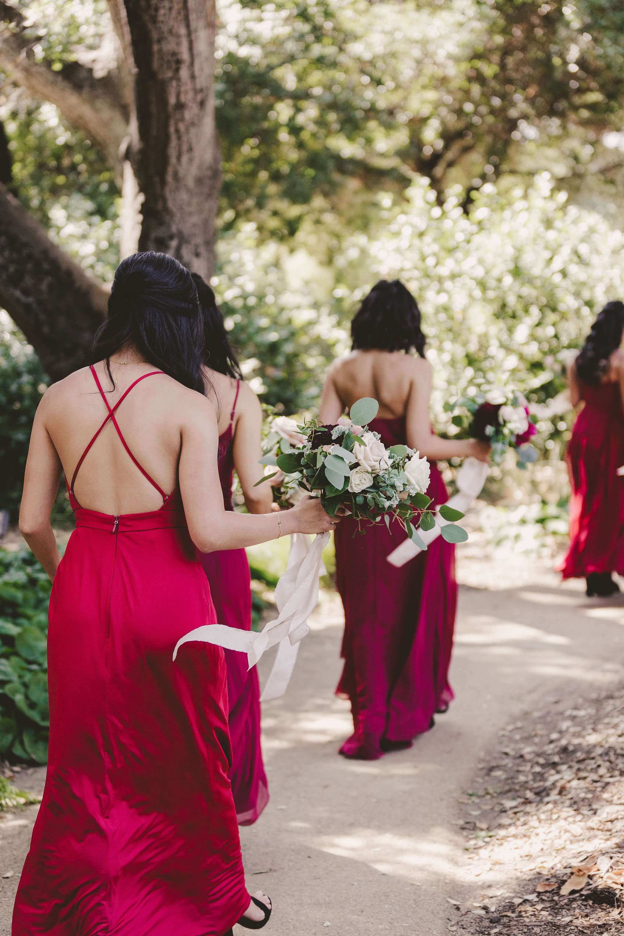 Bridesmaids in red dresses walking on a garden path, holding bouquets with white and pink flowers.
