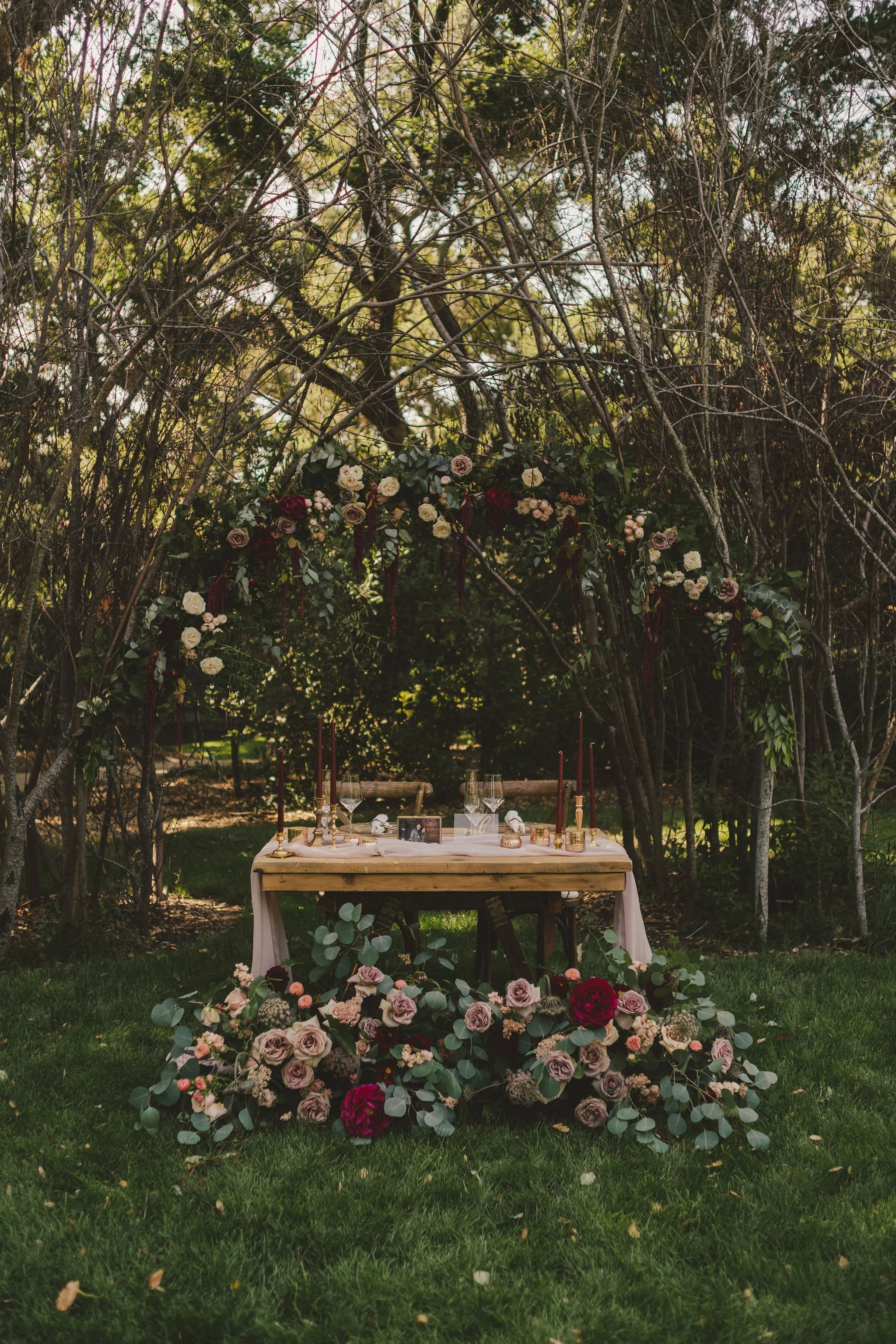 Outdoor wedding table setting with floral arch, candles, and lush greenery.