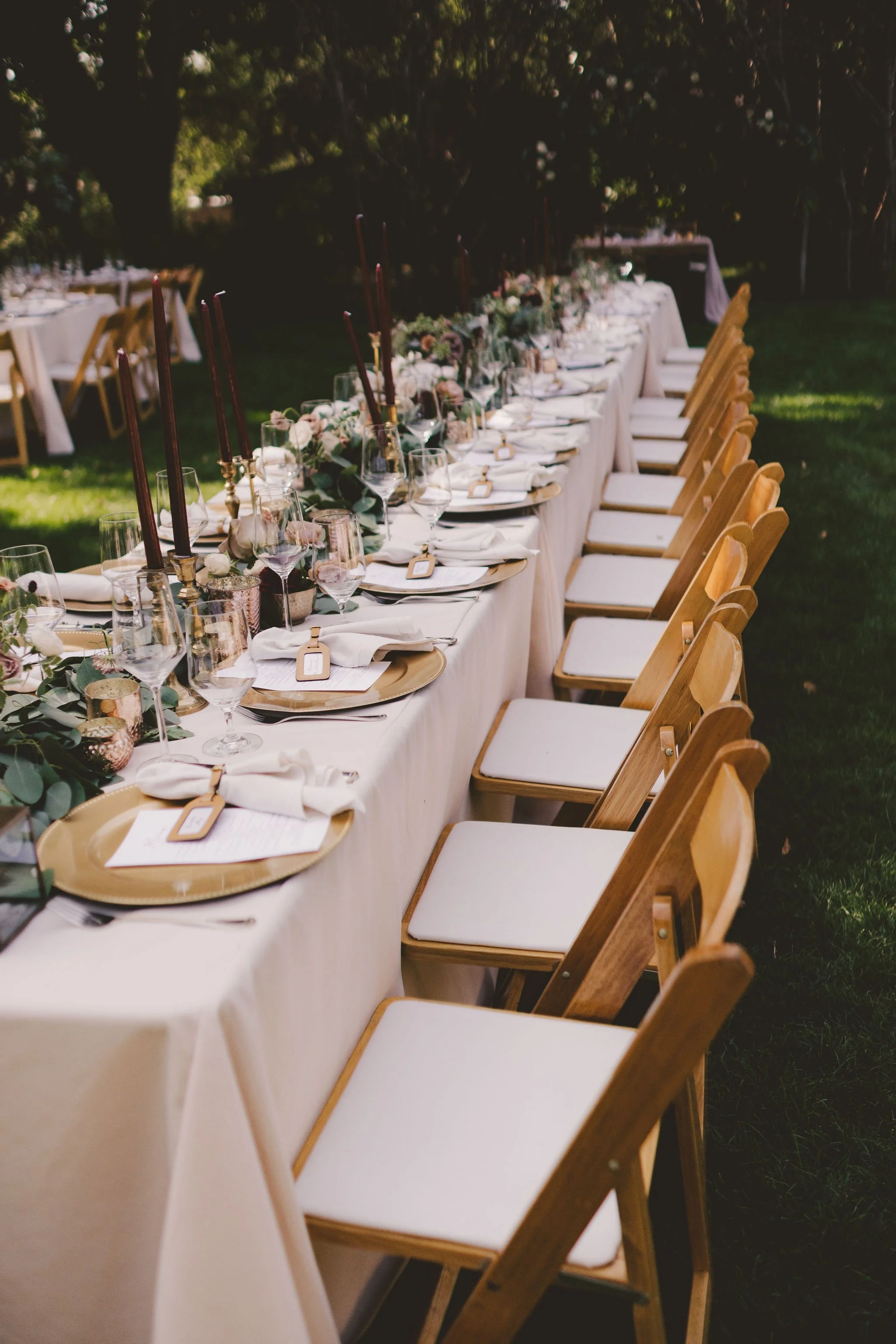 Elegant outdoor banquet table set with white tablecloths, wooden chairs, gold chargers, white napkins, wine glasses, and greenery centerpieces.