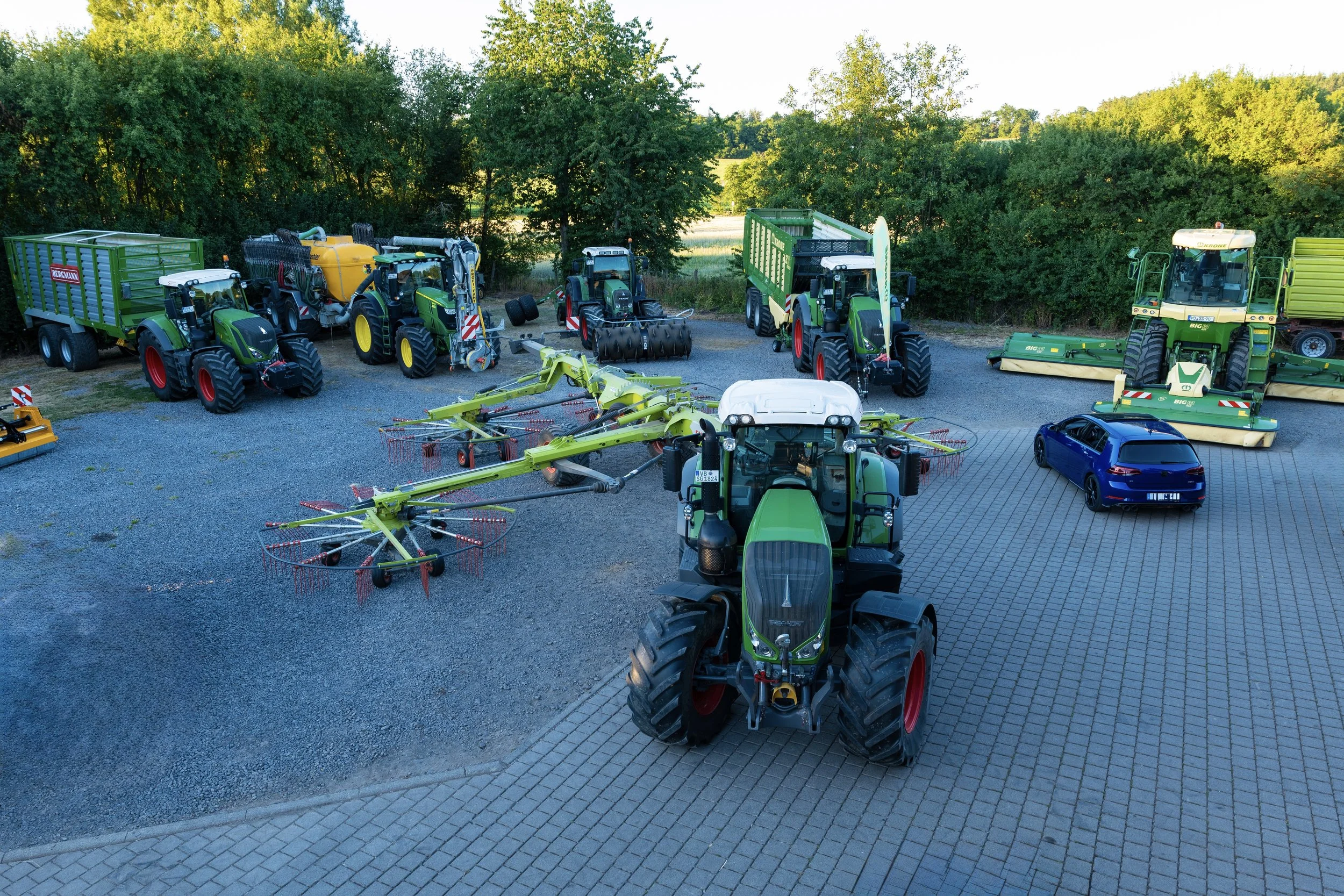 Landwirtschaftliche Maschinen und ein Auto auf einem gepflasterten Platz; Traktoren, Erntemaschinen und Anhänger, umgeben von Bäumen.